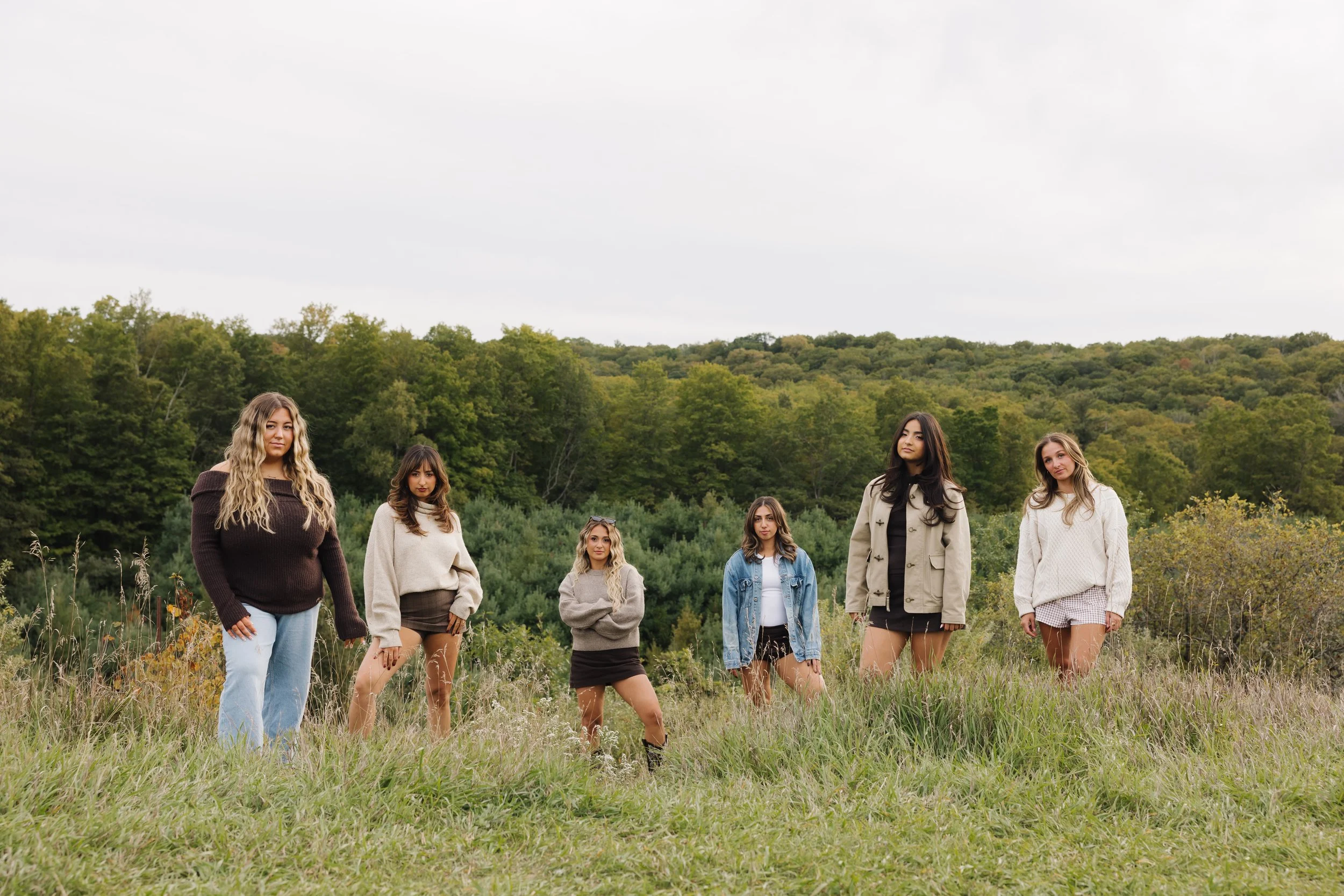Six women posing in a grassy field with a wooded landscape behind them
