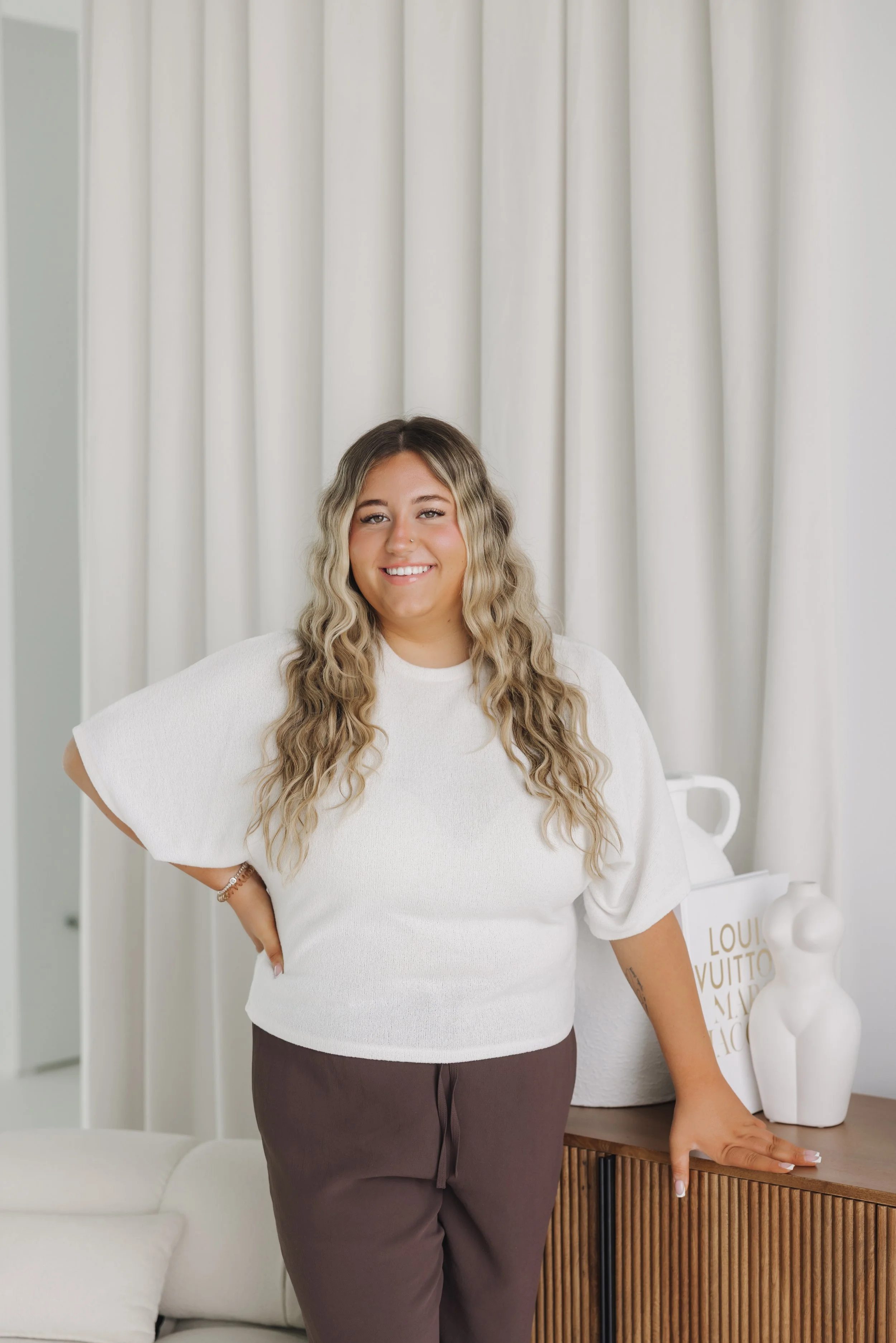Smiling woman with long wavy blonde hair standing in a bright room wearing a white top and brown pants