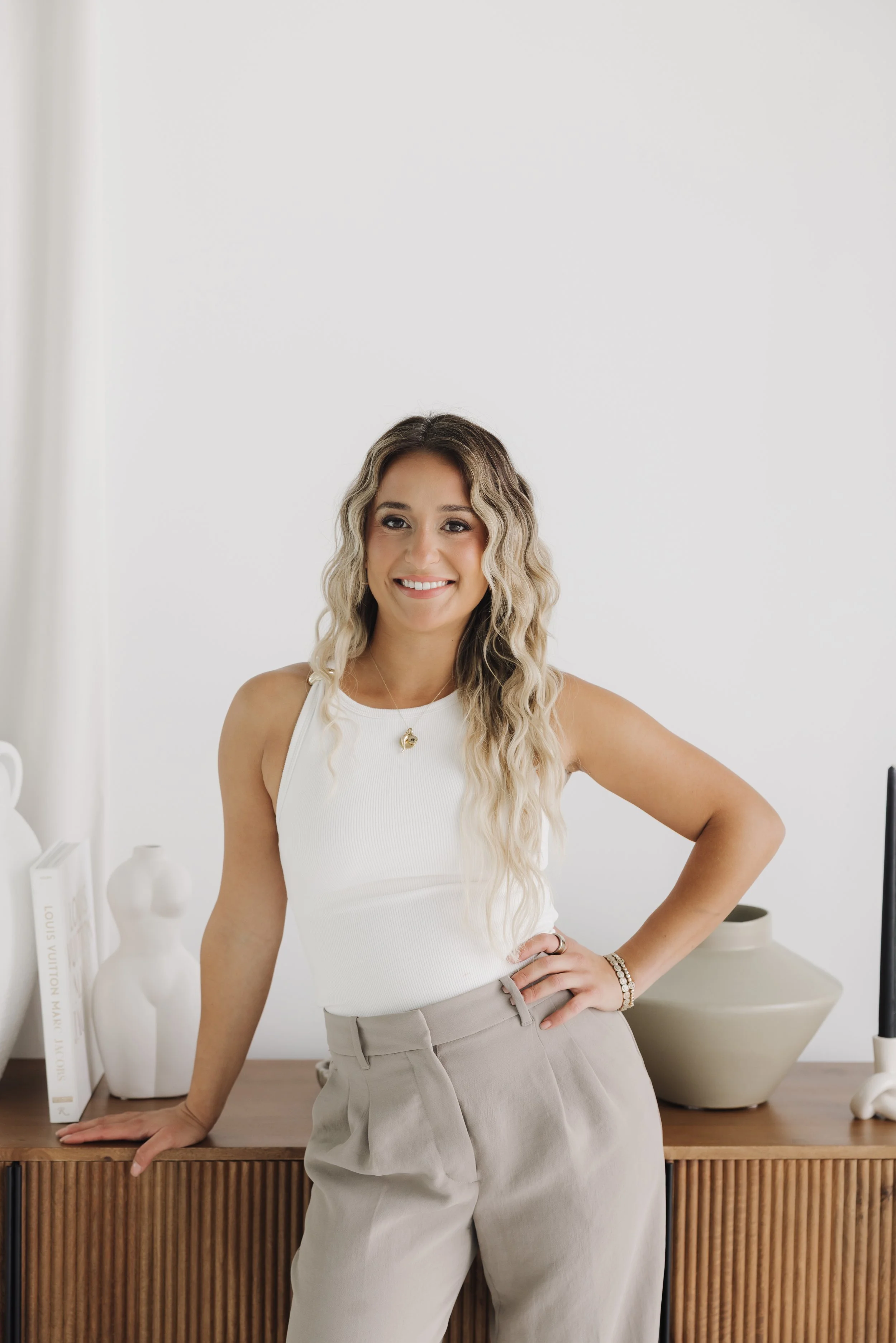Smiling woman with long wavy blonde hair wearing a white sleeveless top and beige pants in a bright home interior