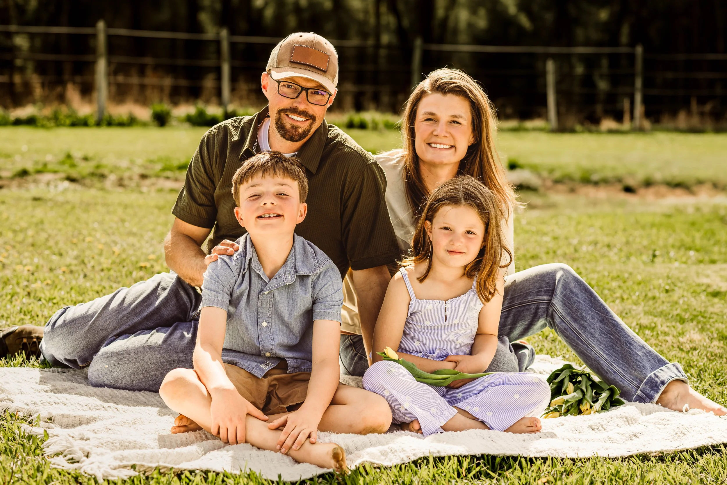 A family of four outside on a sunny day, sitting on a blanket on the grass. The father is wearing glasses and a baseball cap, the mother has long brown hair, and the two children are smiling and sitting close together.