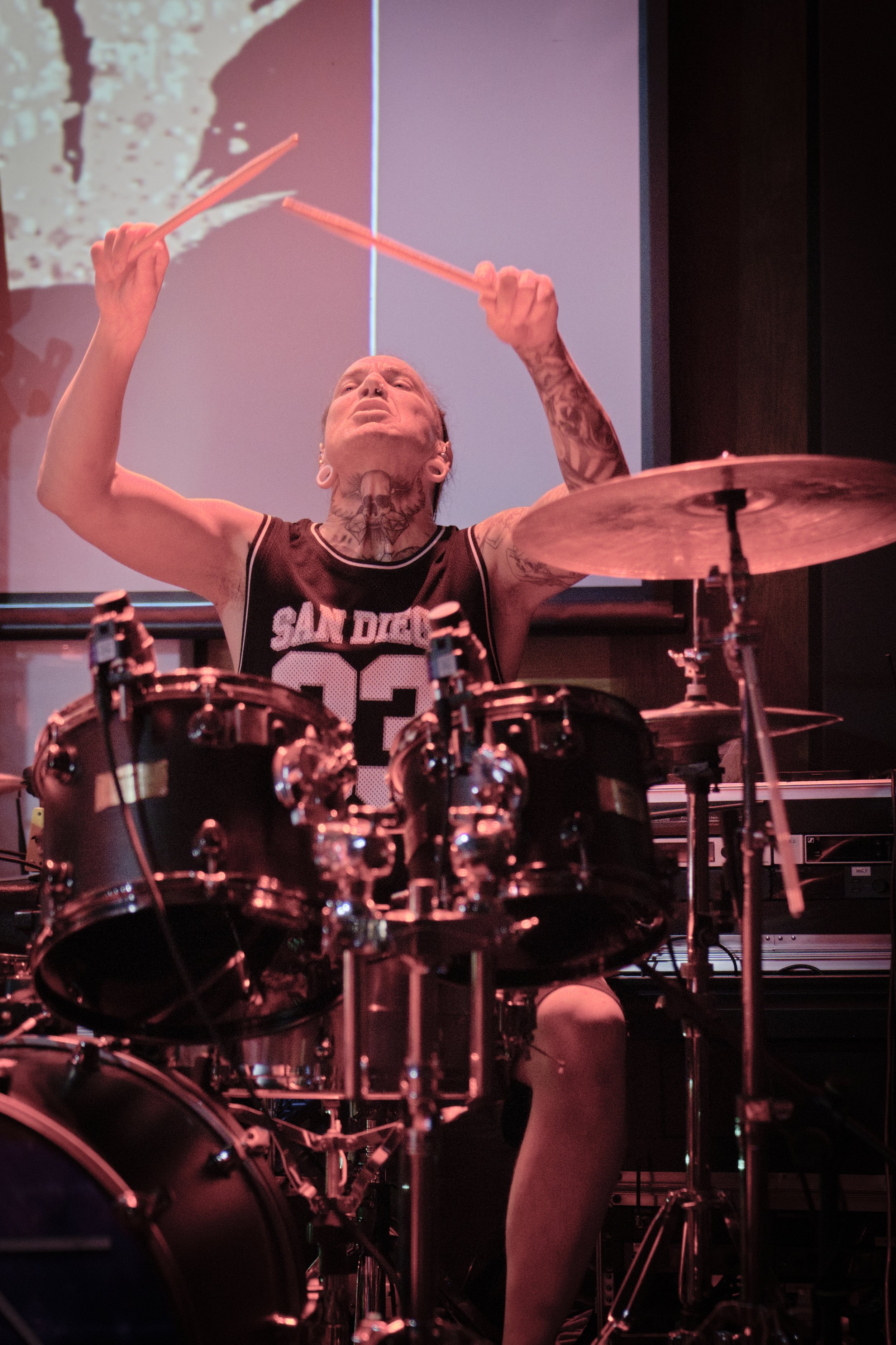 A person with tattoos on their neck and arms playing a drum set and holding drumsticks in the air, wearing a sleeveless sports jersey, in a room with a large screen and dark walls.