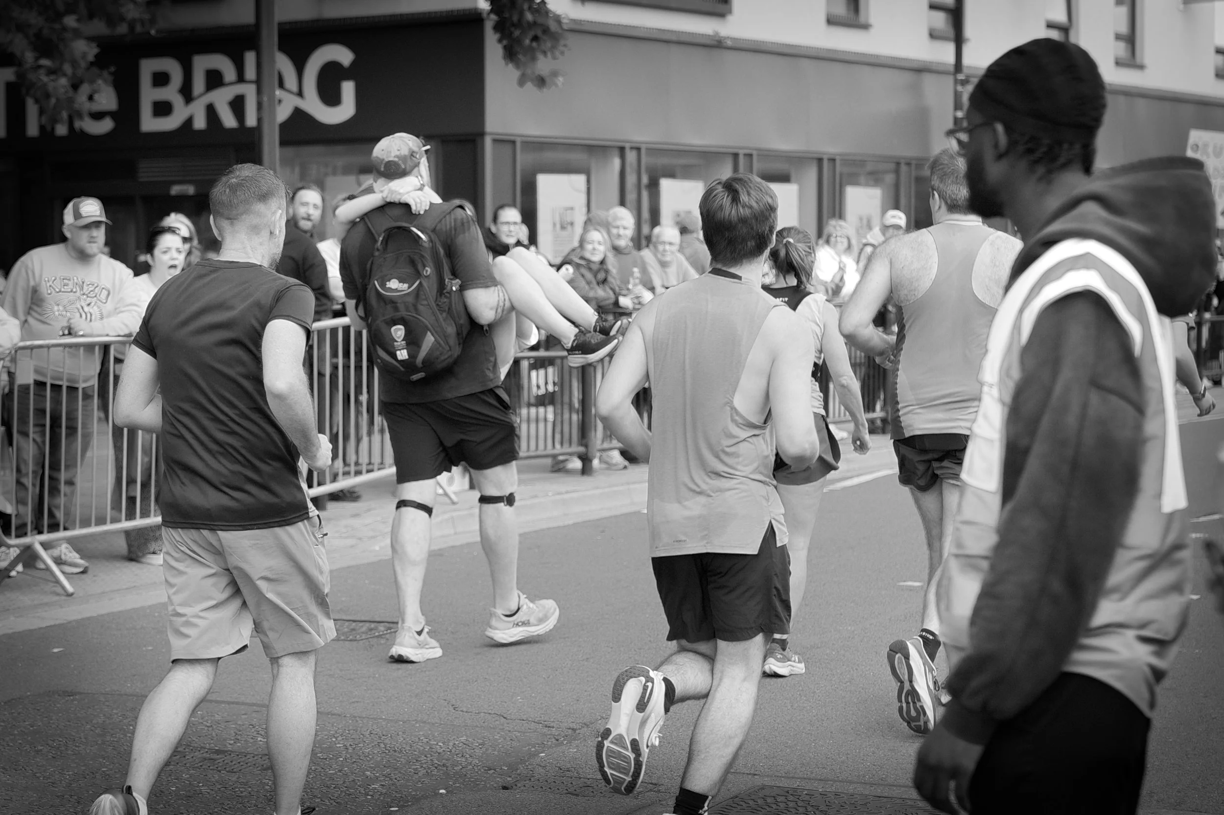 A group of runners participating in a race, with spectators watching behind metal barricades, in an urban setting.