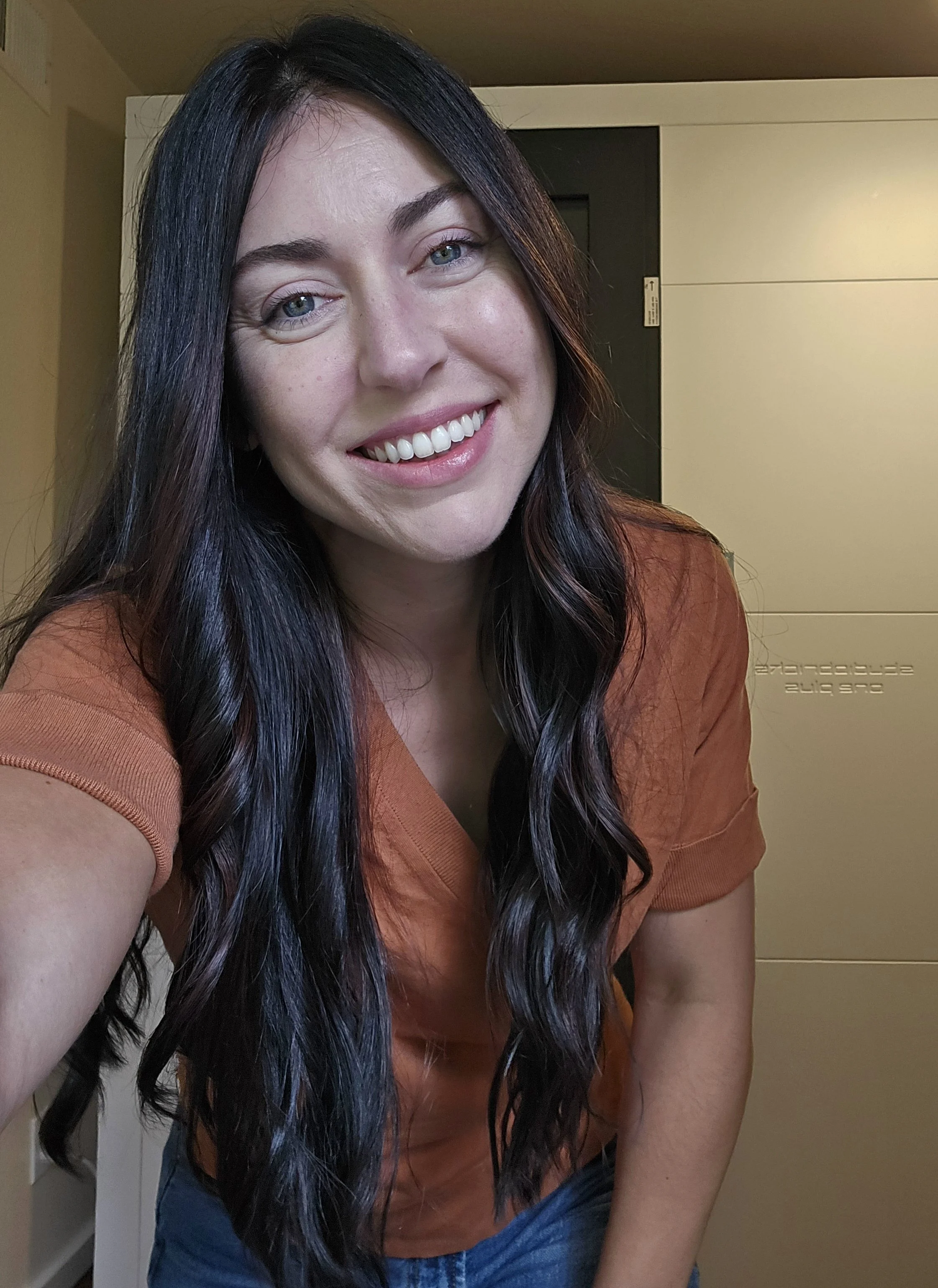 Close-up of a smiling woman with long dark hair with blue streaks, wearing an orange shirt and blue jeans, indoors with a pale yellow wall and a closed dark door in the background.