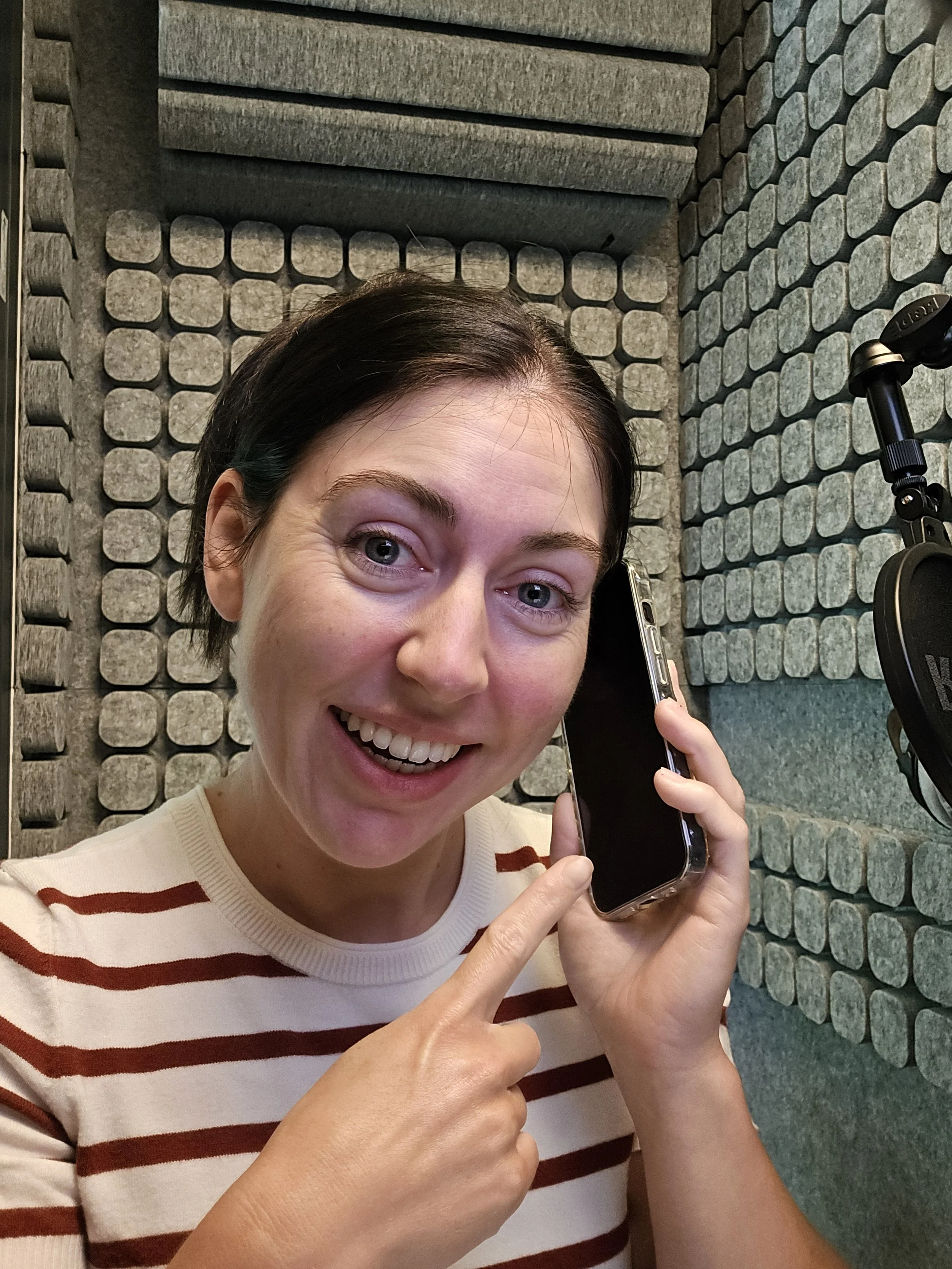 A woman holding a cell phone in front of her face, smiling, inside a voiceover booth with acoustic panels on the walls.