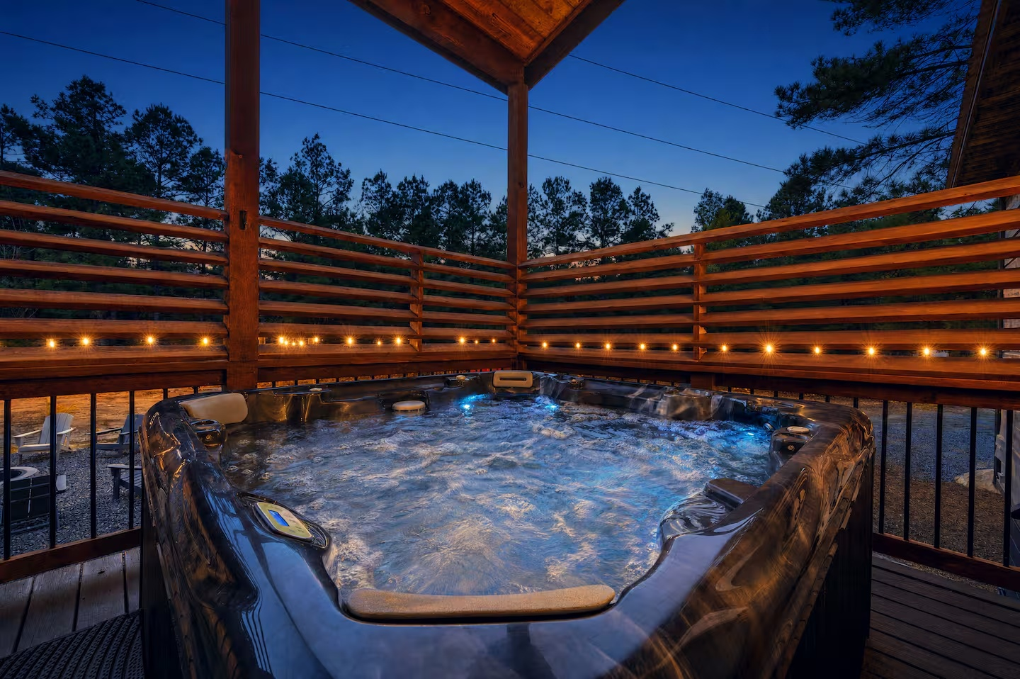 A wooden deck with a hot tub at dusk, surrounded by a wooden railing with string lights, overlooking a forested area with tall trees under a clear blue sky.