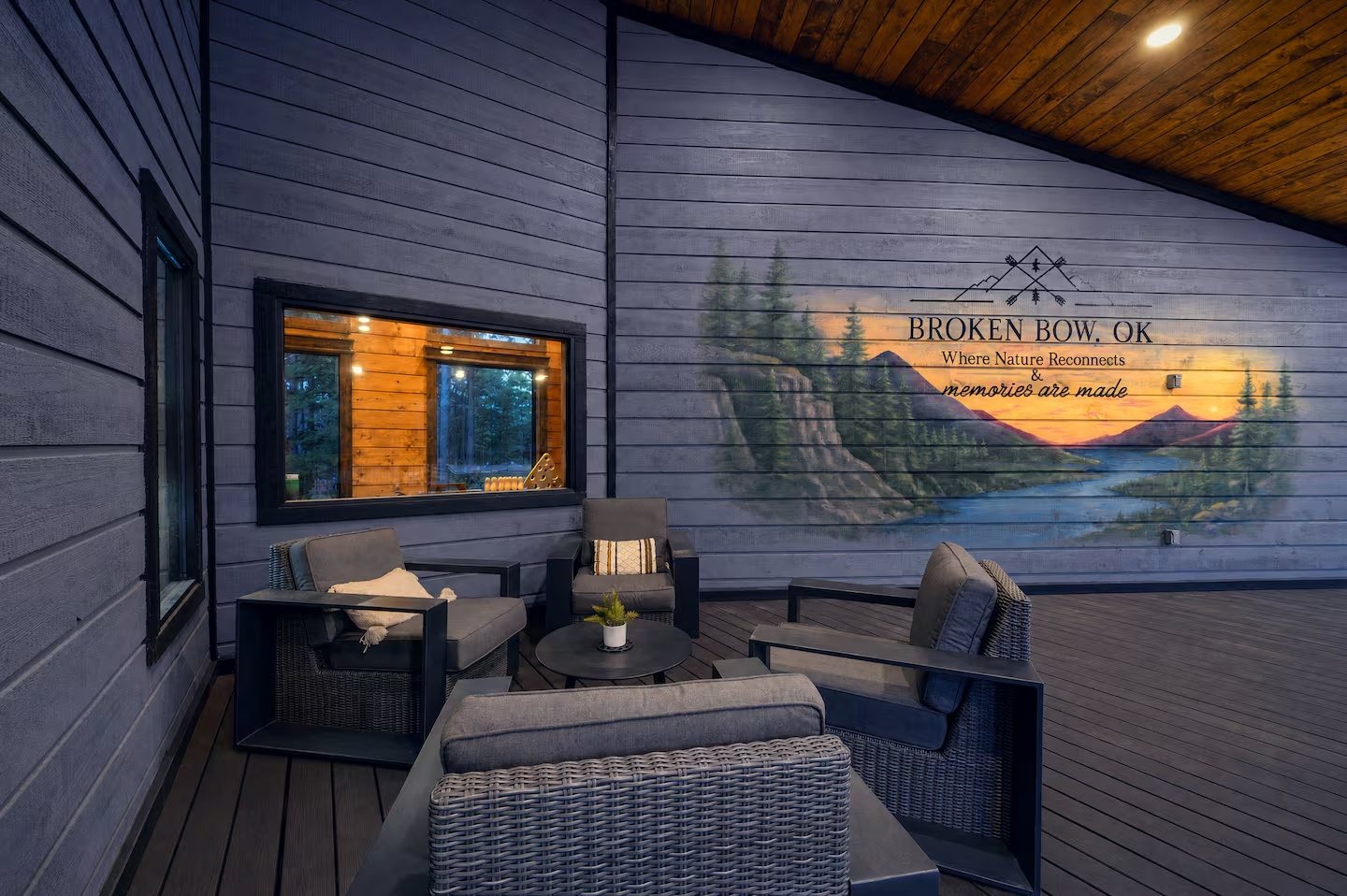Outdoor porch with wicker chairs and a small table, decorated with a potted plant, overlooking a painted mural of mountains, trees, and a sunset in Broken Bow, Oklahoma.