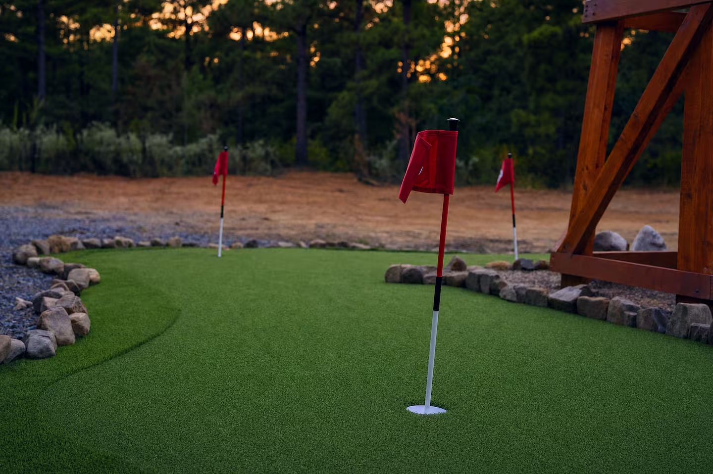 Miniature golf green with flags, surrounded by rocks and a wooden structure, set against a forested background at sunset.