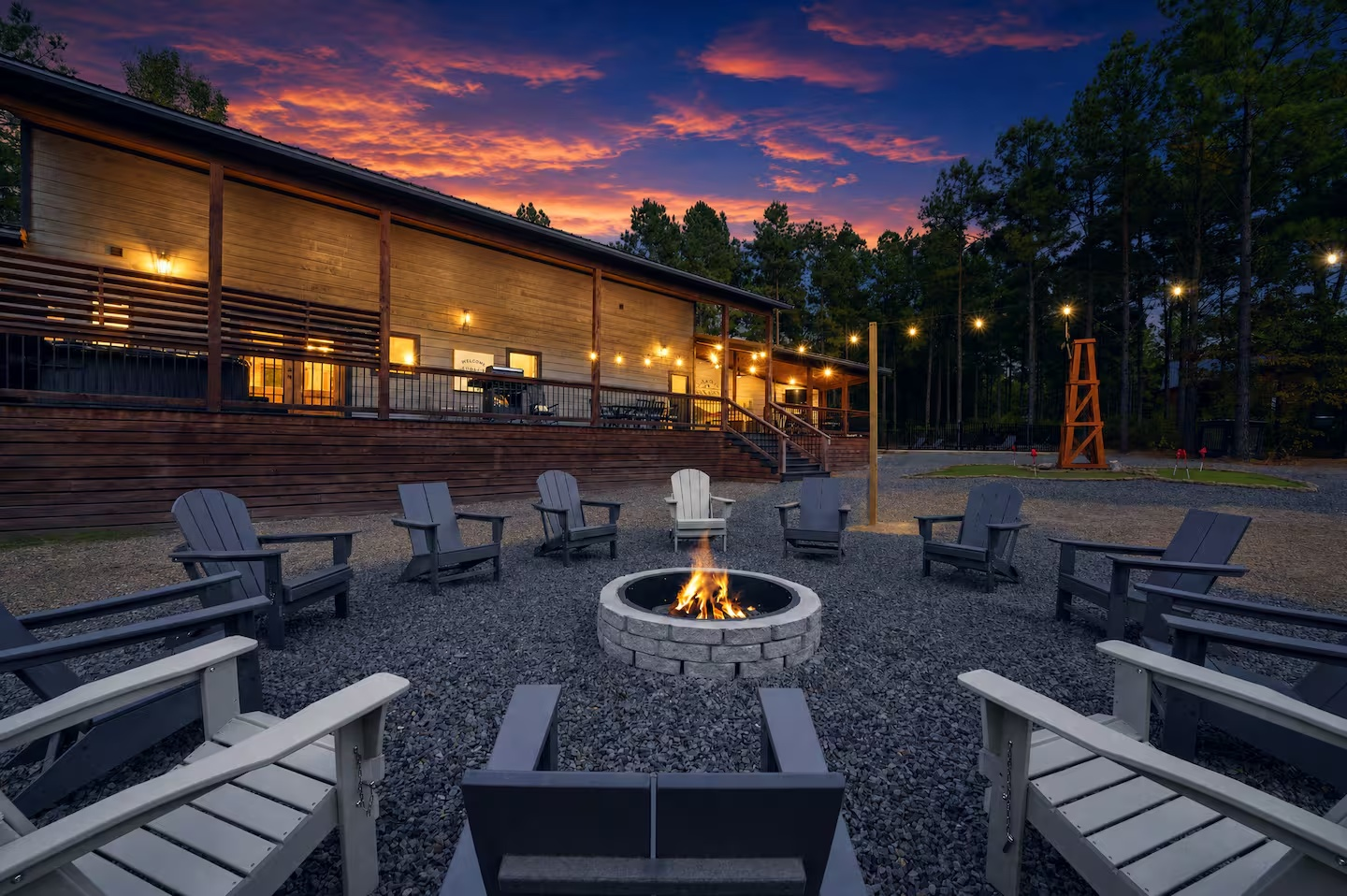 Outdoor patio area at dusk with a fire pit surrounded by Adirondack chairs, a wooden building with string lights, and a forested background with a colorful sunset sky.
