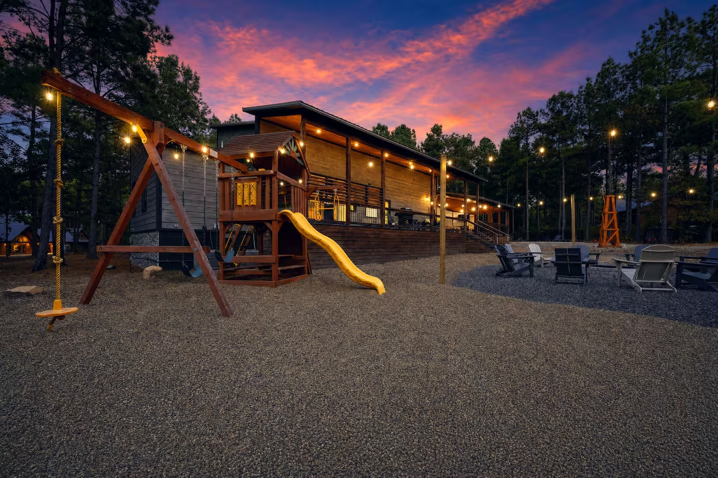 Outdoor area with a wooden house, playground equipment including a swing set and slide, string lights, and chairs at sunset with a colorful sky and trees in the background.