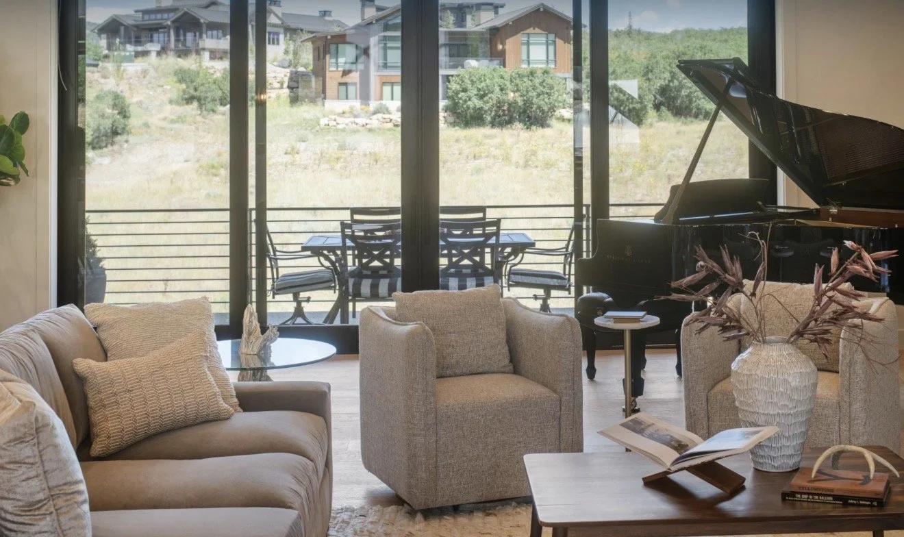 A cozy living room with a beige sofa and two armchairs around a wooden coffee table, which holds a large vase with dried flowers and some books. In the background, there is a black grand piano and a large sliding glass door leading to a balcony with outdoor seating, overlooking a hillside with modern houses and greenery.