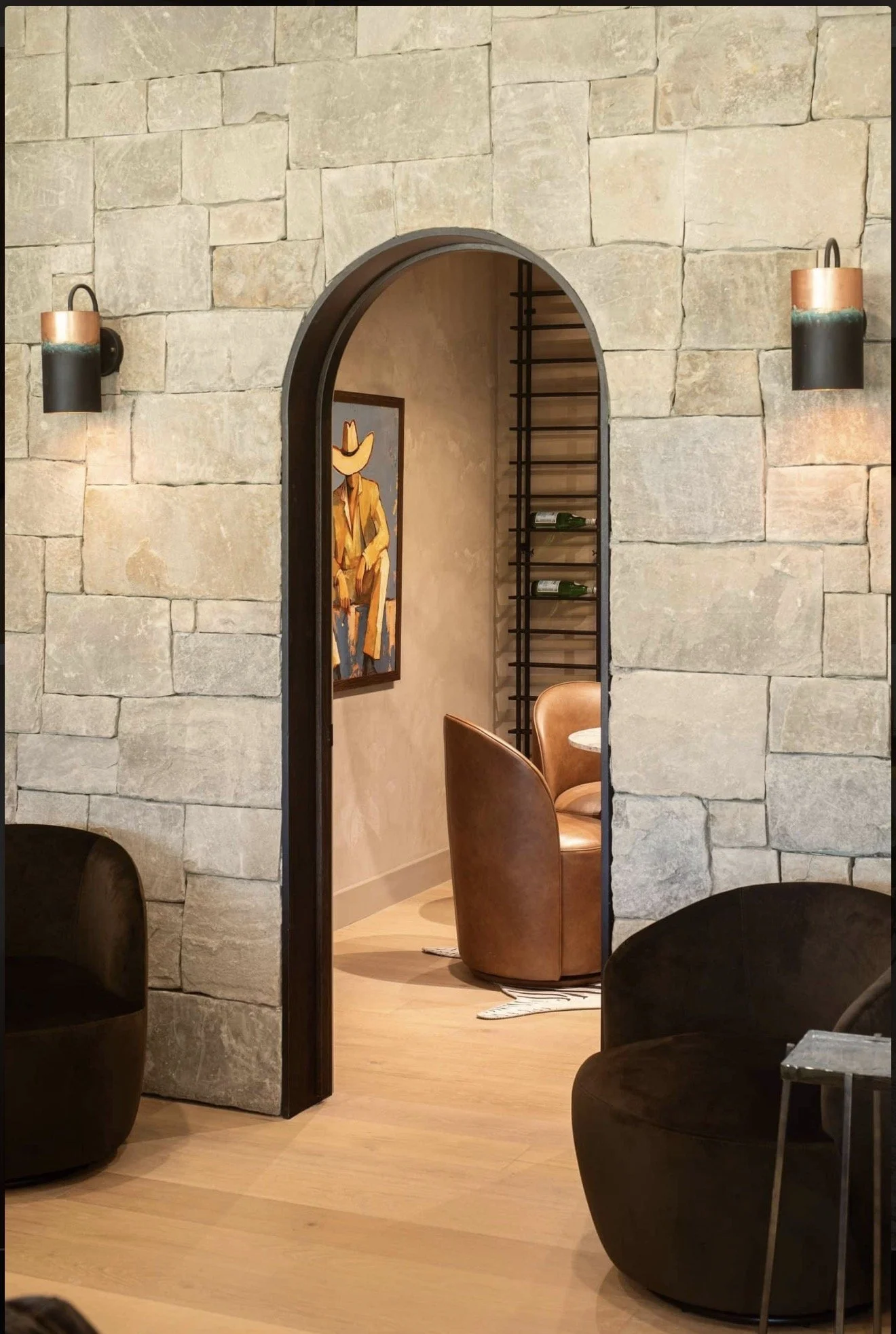 A cozy restaurant interior with stone walls, modern lighting, and artwork of a cowboy. The room features leather chairs and a small table, with a view through an arched doorway into a space with a wall-mounted wine rack and a chair.