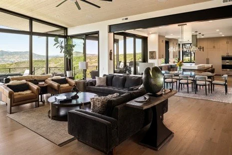 Modern living room with large windows, plush sofas, coffee table, and a view of greenery outside.