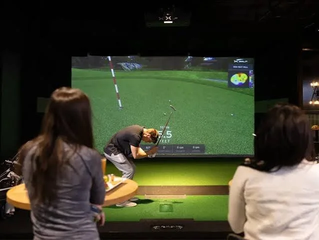 People playing indoor golf simulation game with a large screen displaying a virtual golf course.