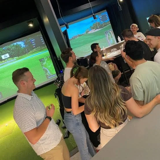 Group of people at a golf simulation bar, some playing virtual golf on large screens, others socializing, with drinks on the bar counter.