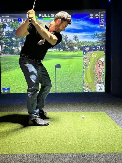 Man practicing golf swing indoors on a virtual golf simulator, with a large screen displaying a golf course background and a side map showing course details.
