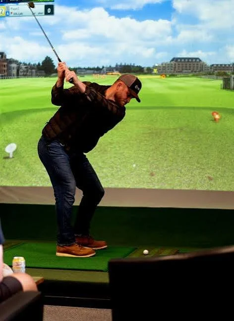 Man in black jacket and cap preparing to hit a golf ball at an indoor golf simulation center with a virtual golf course background.