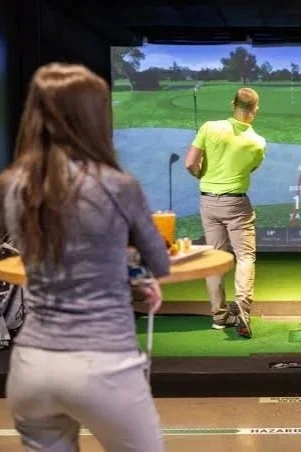 People at an indoor golf simulator, one person watching and another swinging a golf club on the screen.