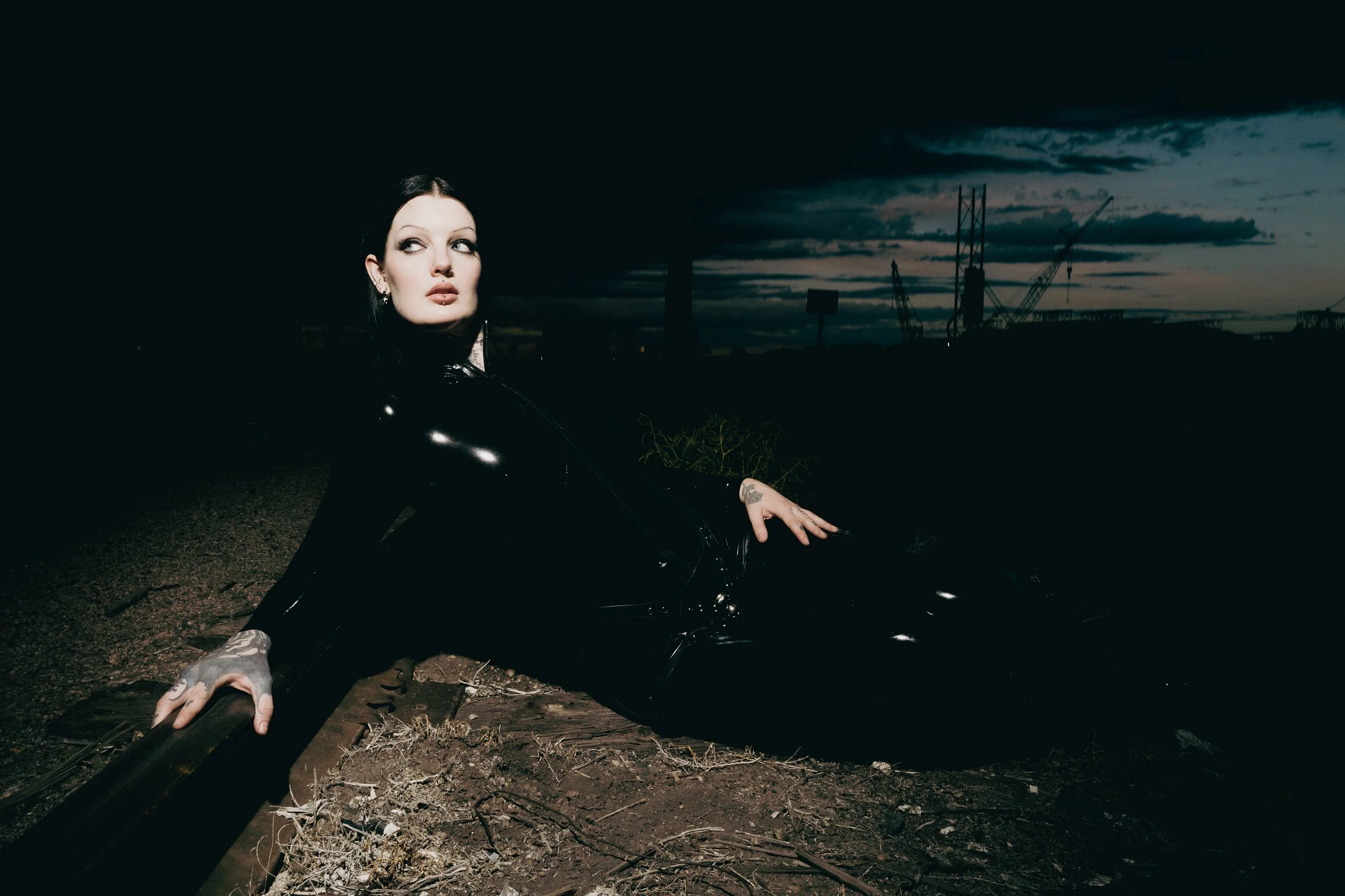 A woman with dark hair and tattoos on her arm, wearing dark makeup and a black outfit, lying on a railway track at dusk with a dark cloudy sky and construction cranes in the background.