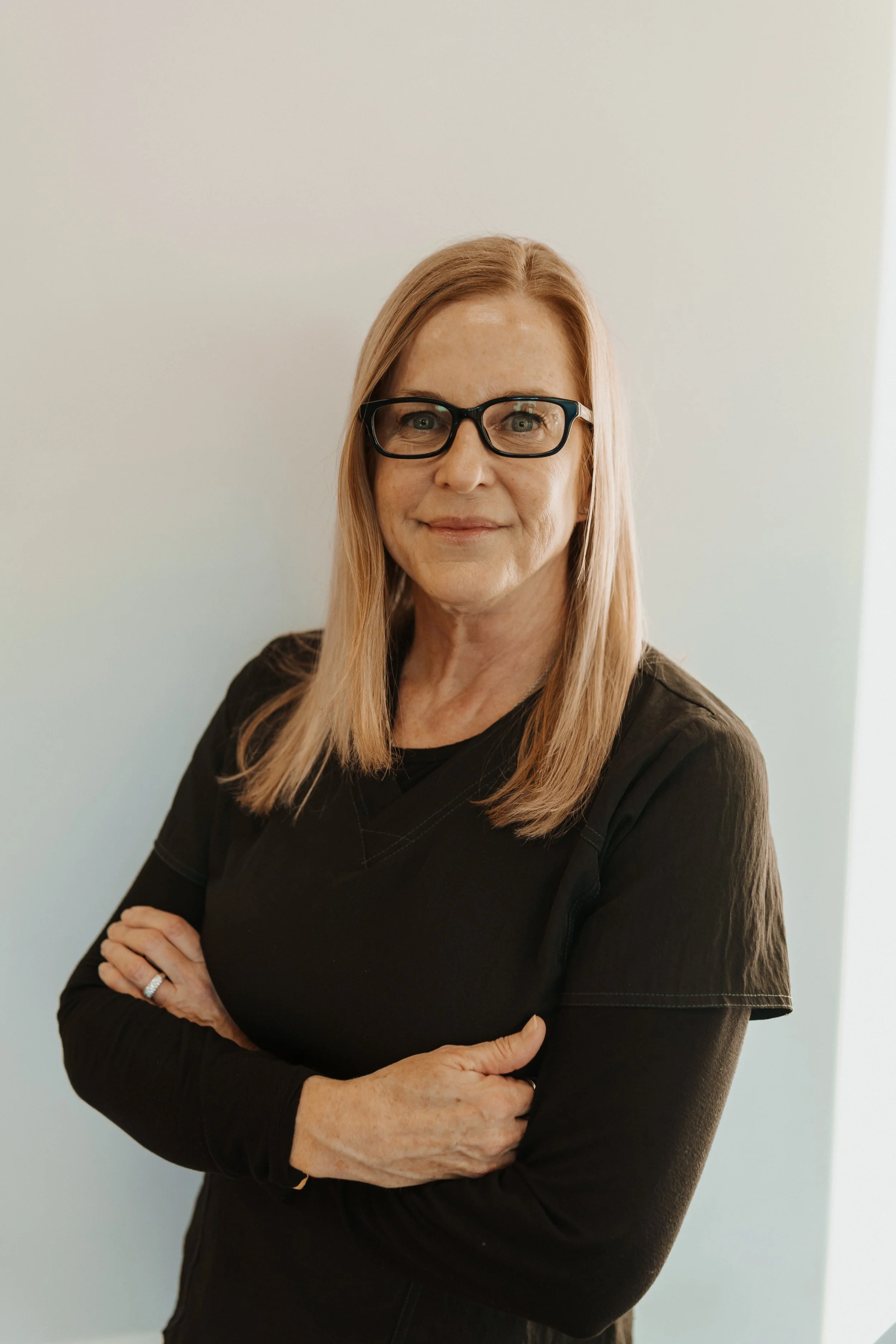 Portrait of a smiling mature woman with glasses and long blonde hair, wearing black and standing with her arms crossed.