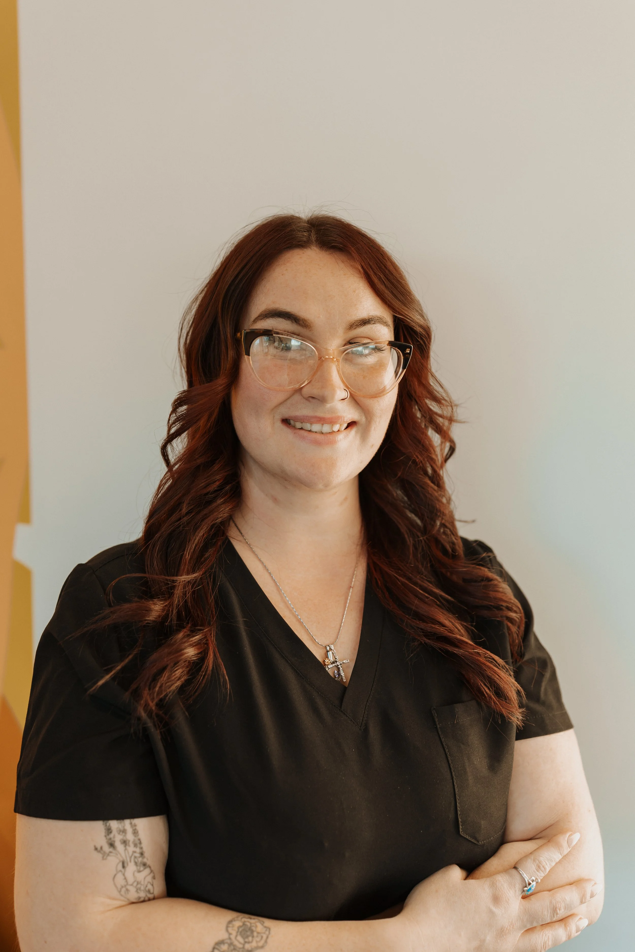 Smiling woman with red hair and glasses wearing black scrubs and a cross necklace