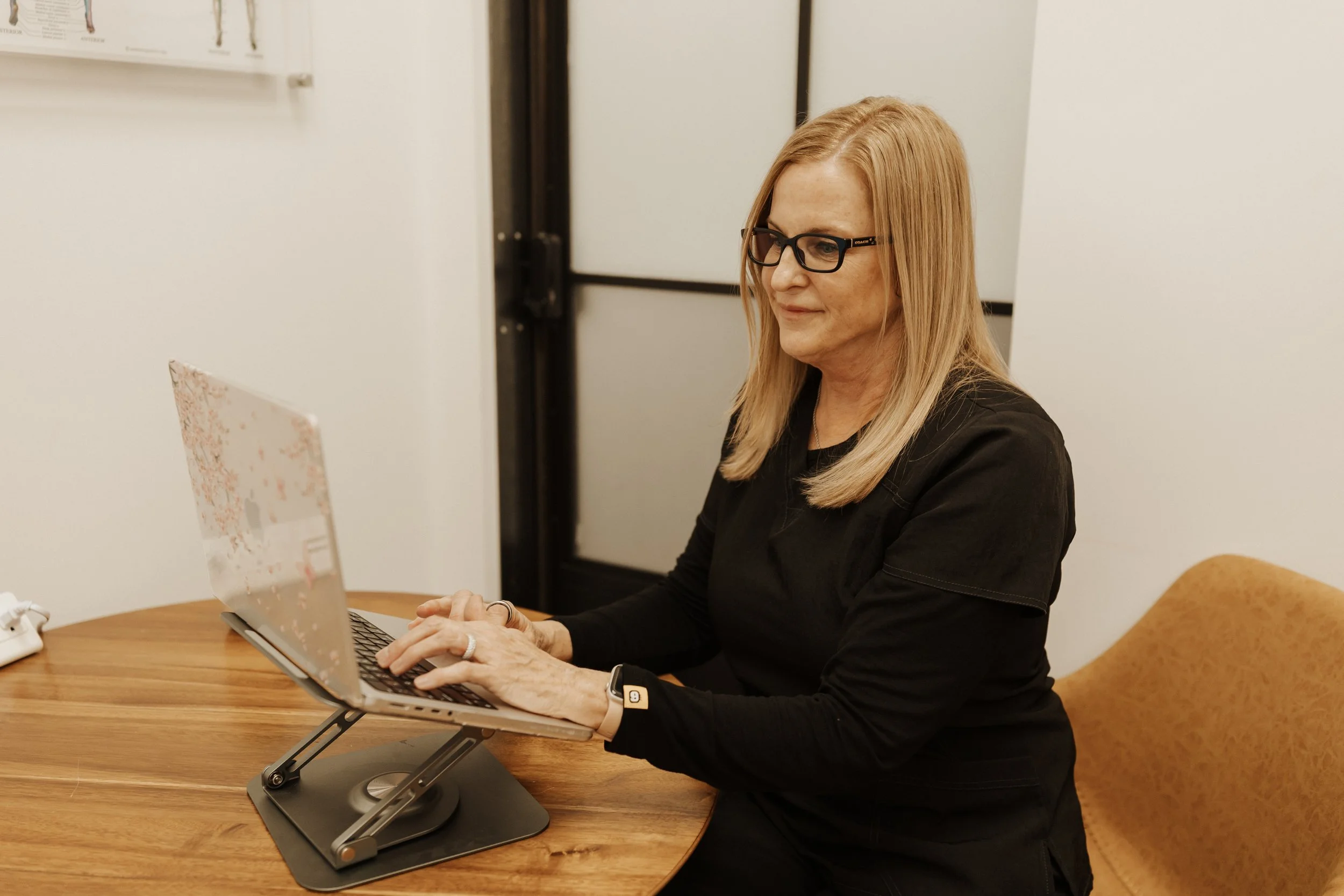 Woman wearing glasses works on a laptop at a wooden desk in an office.