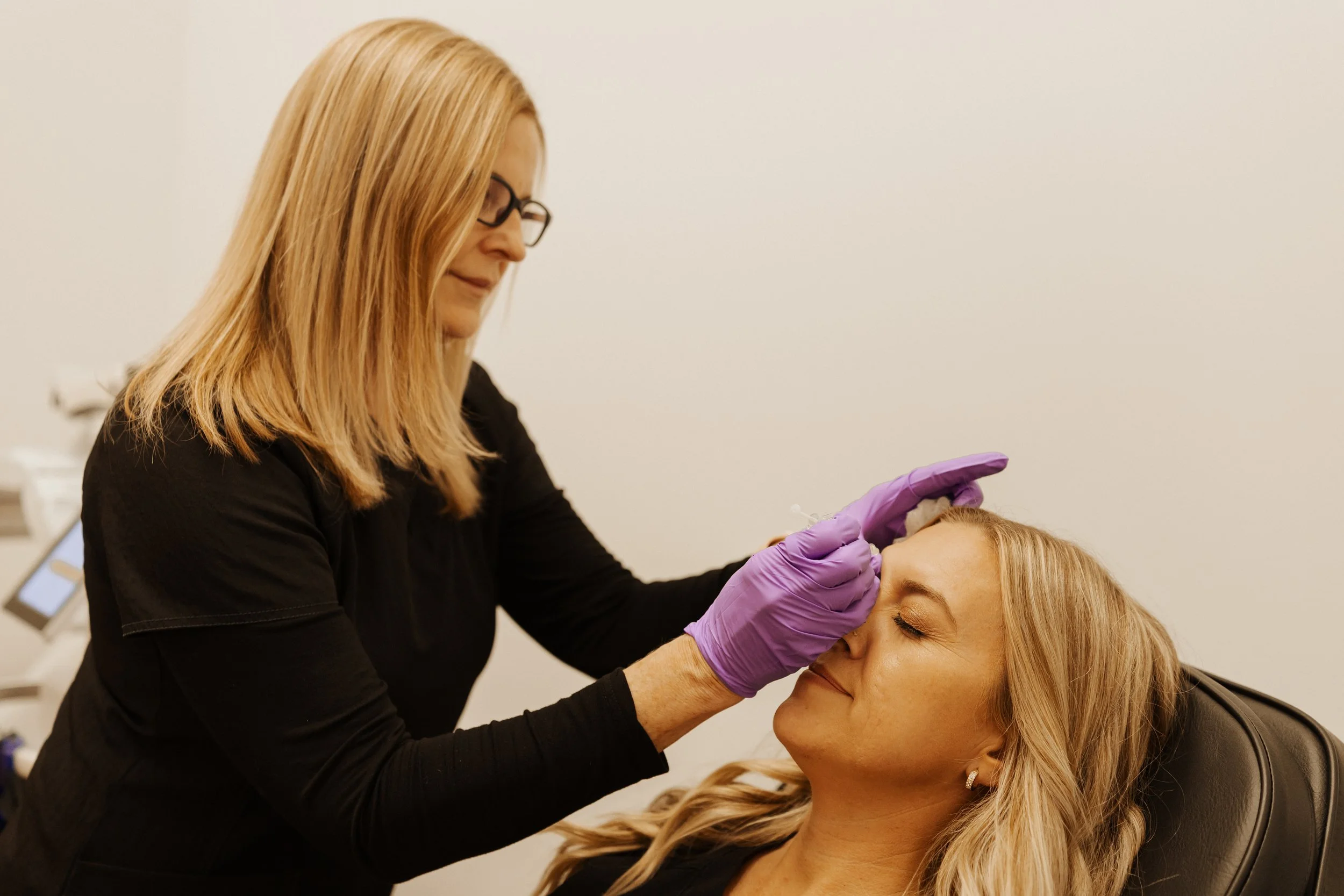 Aesthetician in purple gloves performing a facial treatment on a reclining woman in a spa.