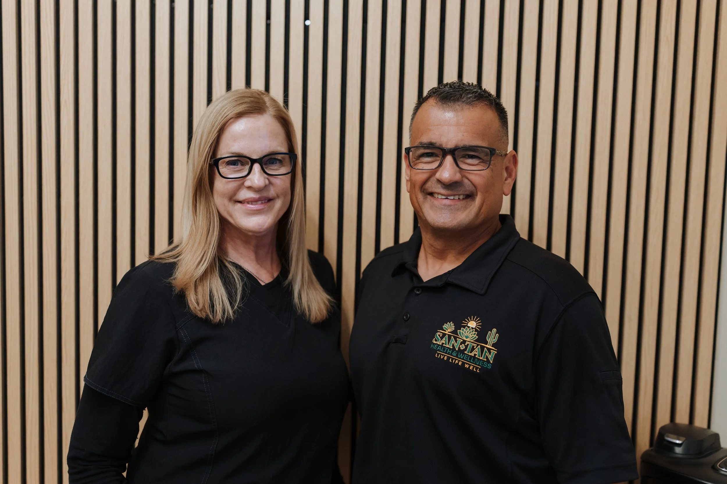 Smiling man and woman in black wellness uniforms standing in front of a wood slat wall
