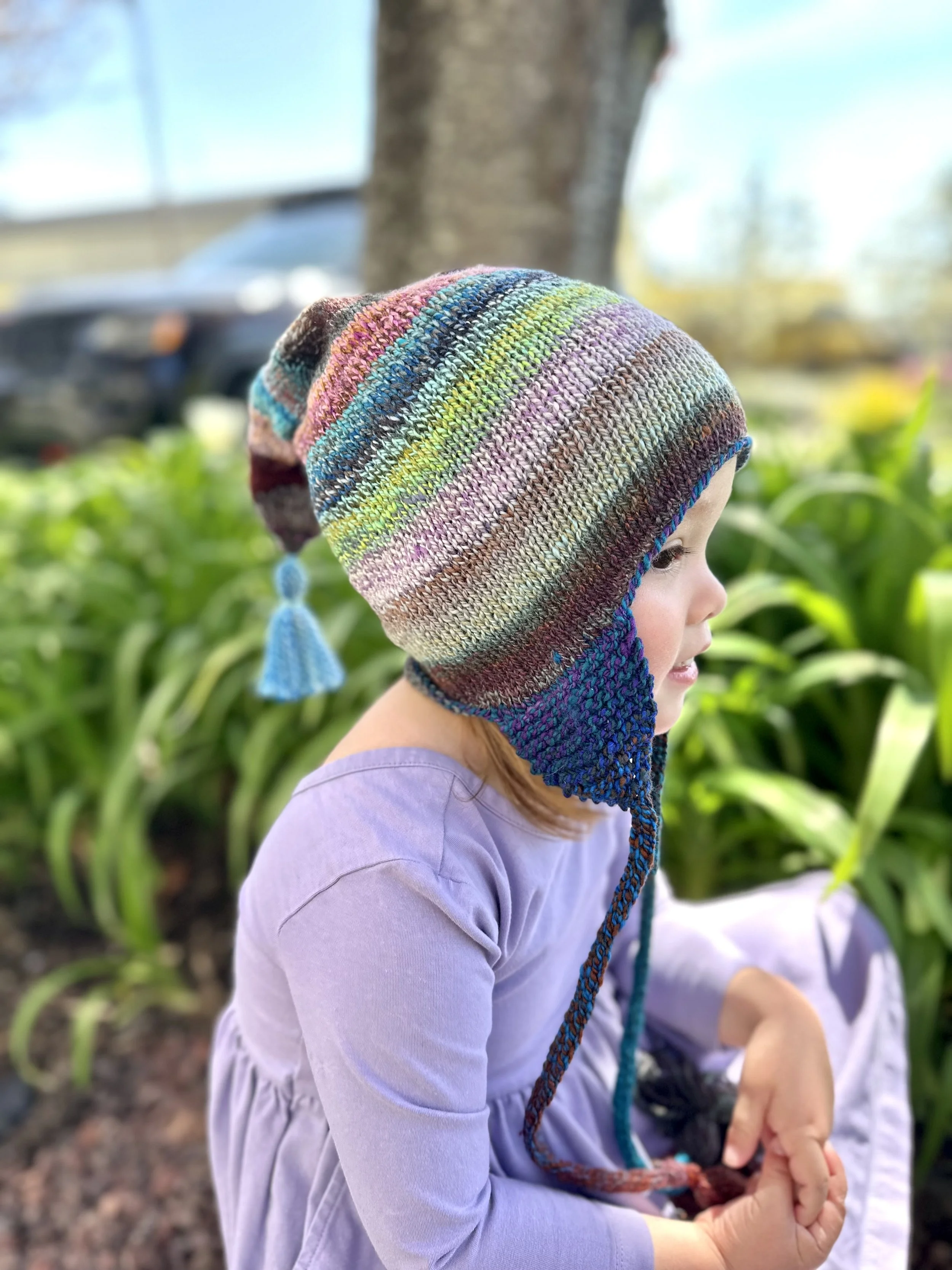 Side view of a child wearing a multicolored Hudson Hat, showing the knitted ear flap, tapered crown and tassel.