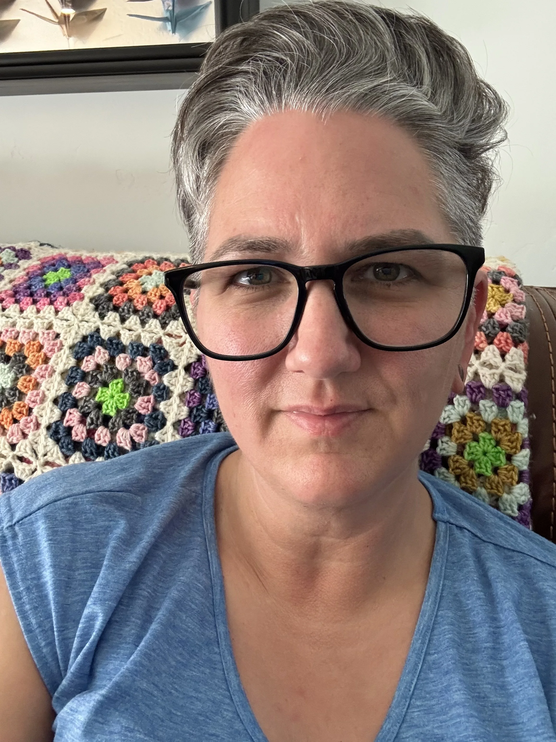 Lindsay Baker wearing black glasses and a blue shirt, seated in front of a colorful crocheted blanket.