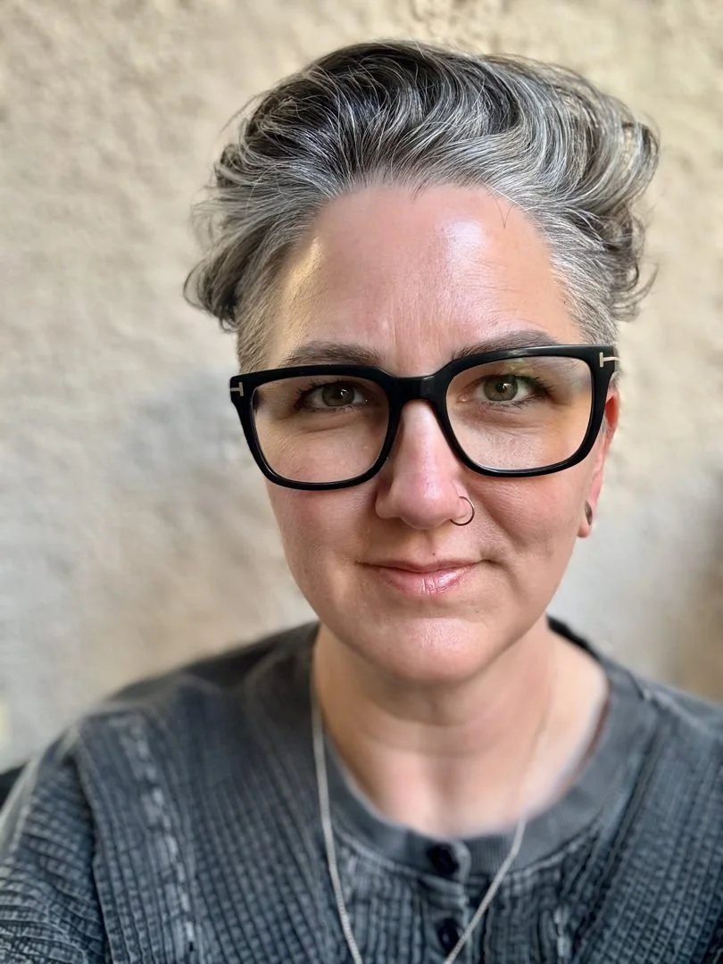 Lindsay wearing black glasses and a gray sweater against a softly textured background.