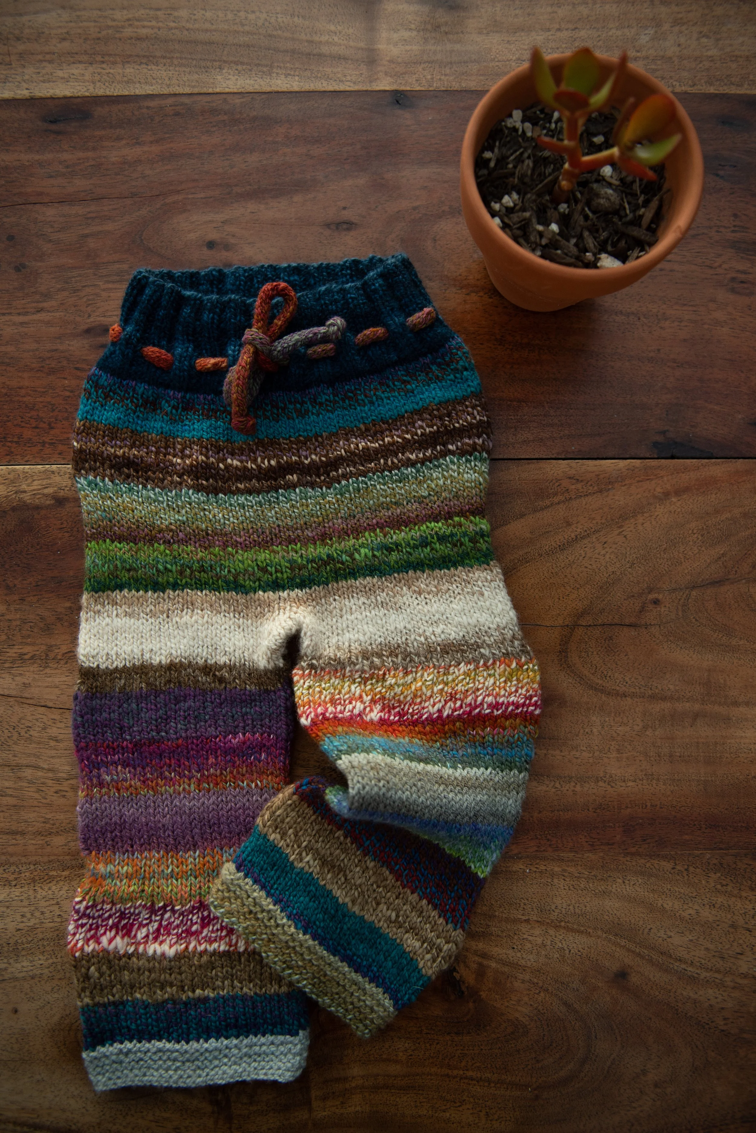 Striped knitted baby pants with a dark teal ribbed waistband, i-cord drawstring and garter-stitch cuffs, laid on a wooden table.