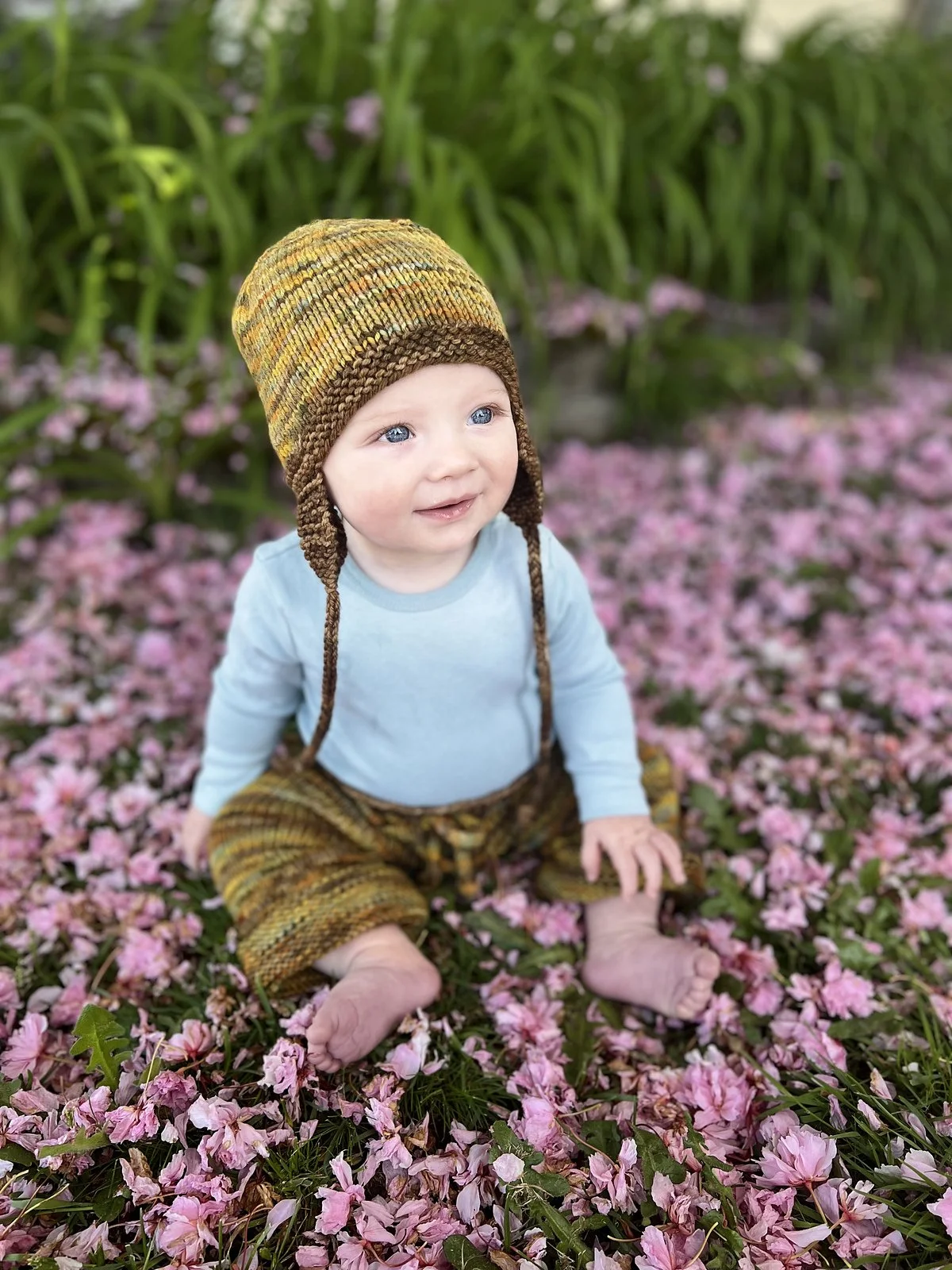 Baby wearing a gold and brown knitted ear-flap hat with i-cord ties, seated among fallen pink petals.