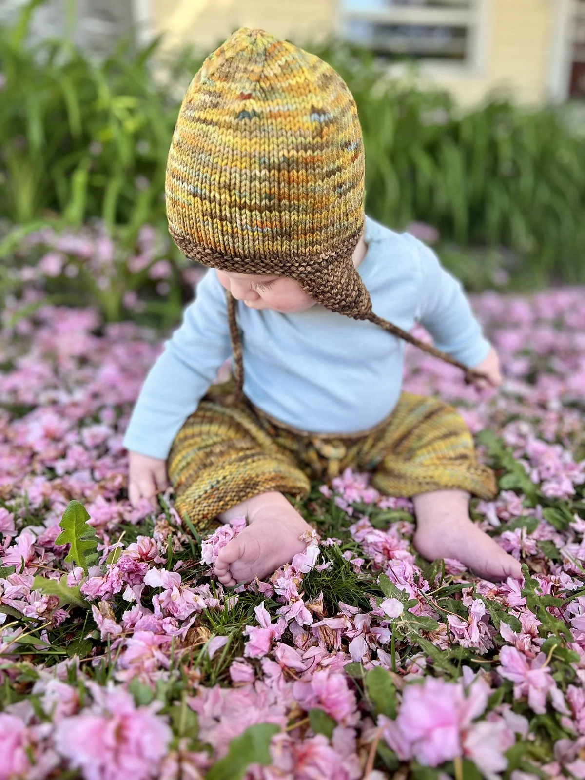 View of the shaped crown and garter-stitch ear flaps of the Ky Baby hat, worn by a baby in a garden.