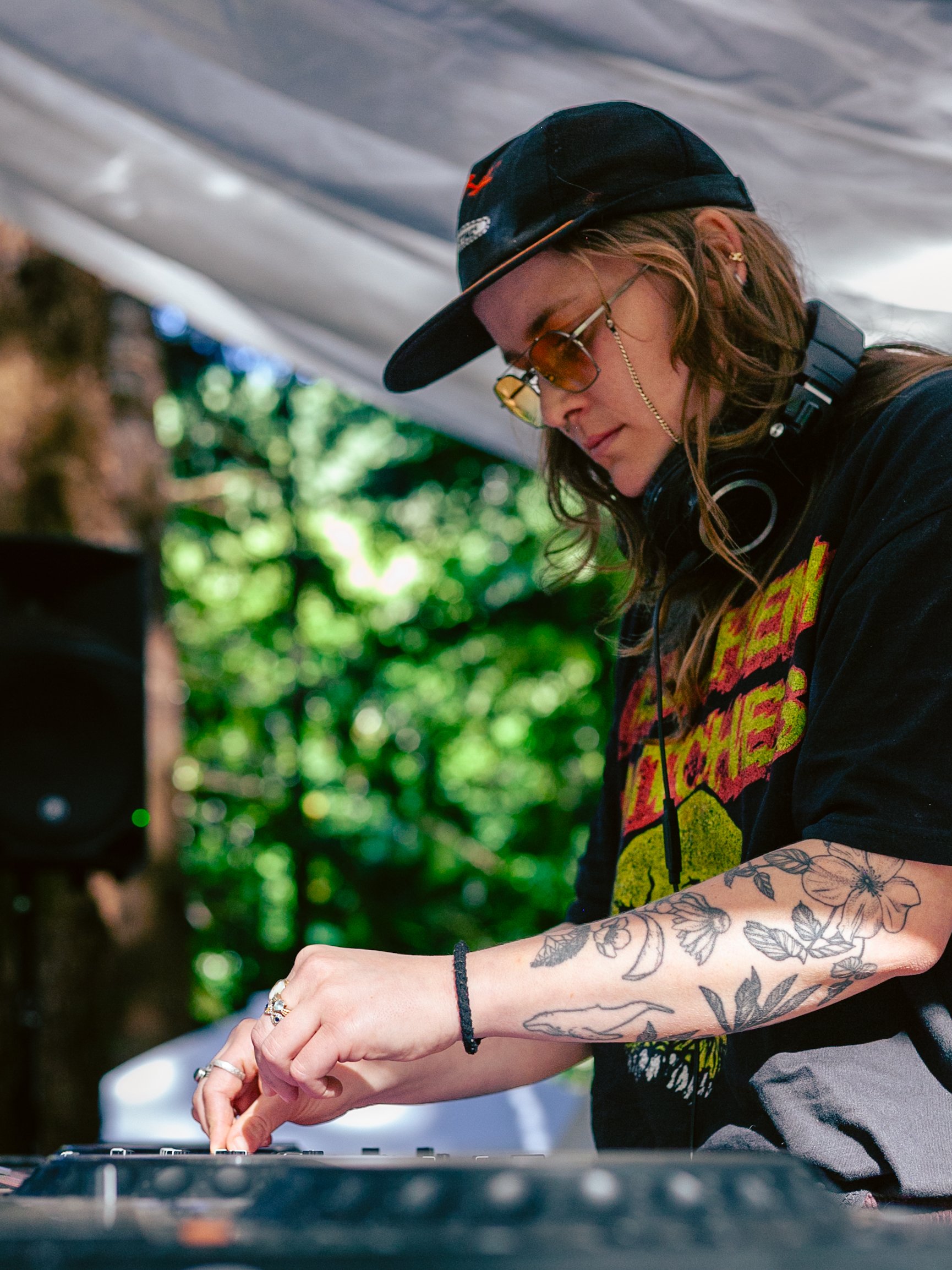 A person wearing sunglasses, a black cap, and floral tattoos on their arm is DJing at an outdoor event with green foliage in the background.