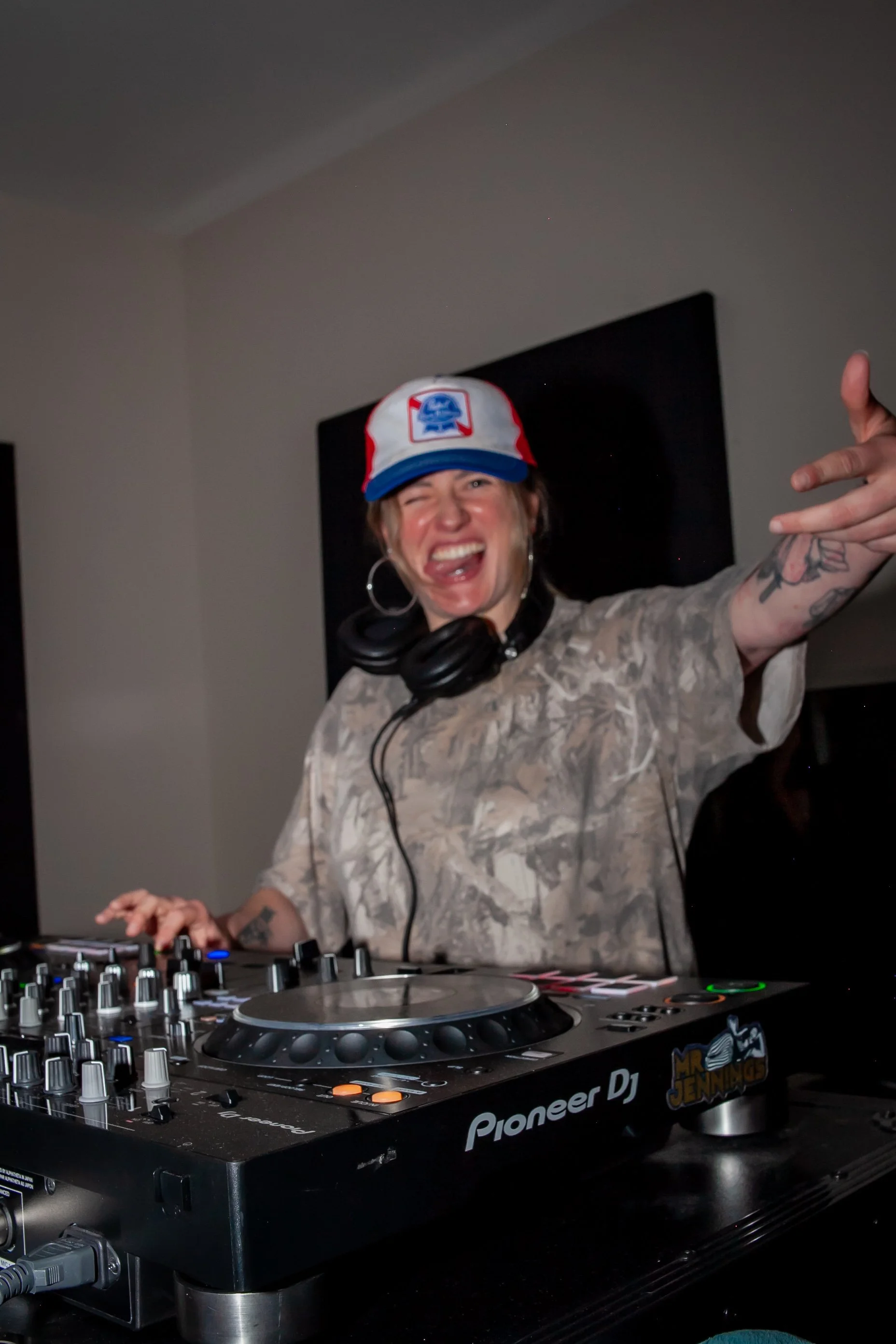 A woman with a baseball cap, large hoop earrings, and tattoos on her arm is DJing with headphones around her neck, smiling and pointing in the air, at a DJ deck with Pioneer DJ equipment.