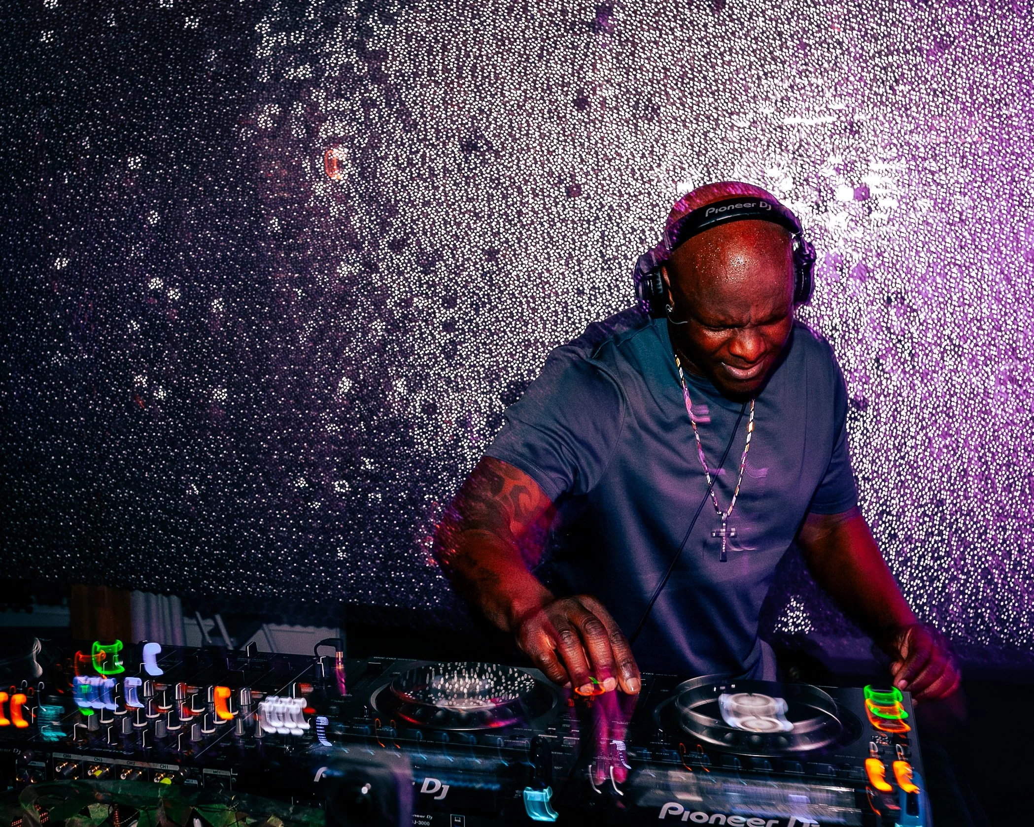 A DJ with a shaved head wearing headphones, playing music at a vibrant nightclub with colorful lighting and a glittery backdrop.