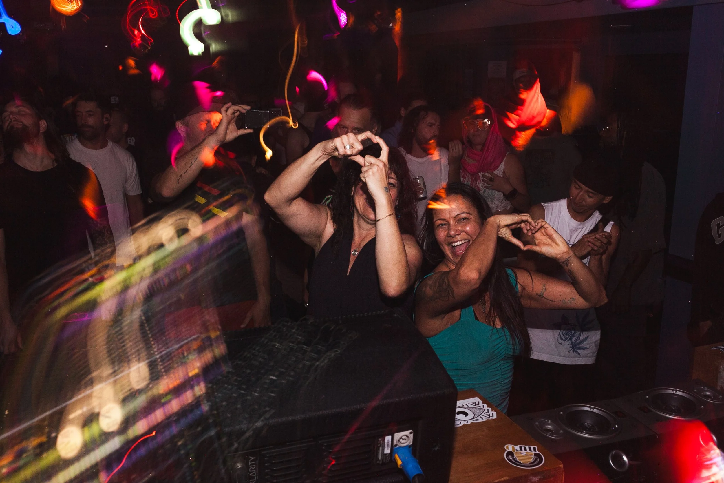People dancing and having fun in a nightclub with colorful lights and a DJ booth in the foreground.