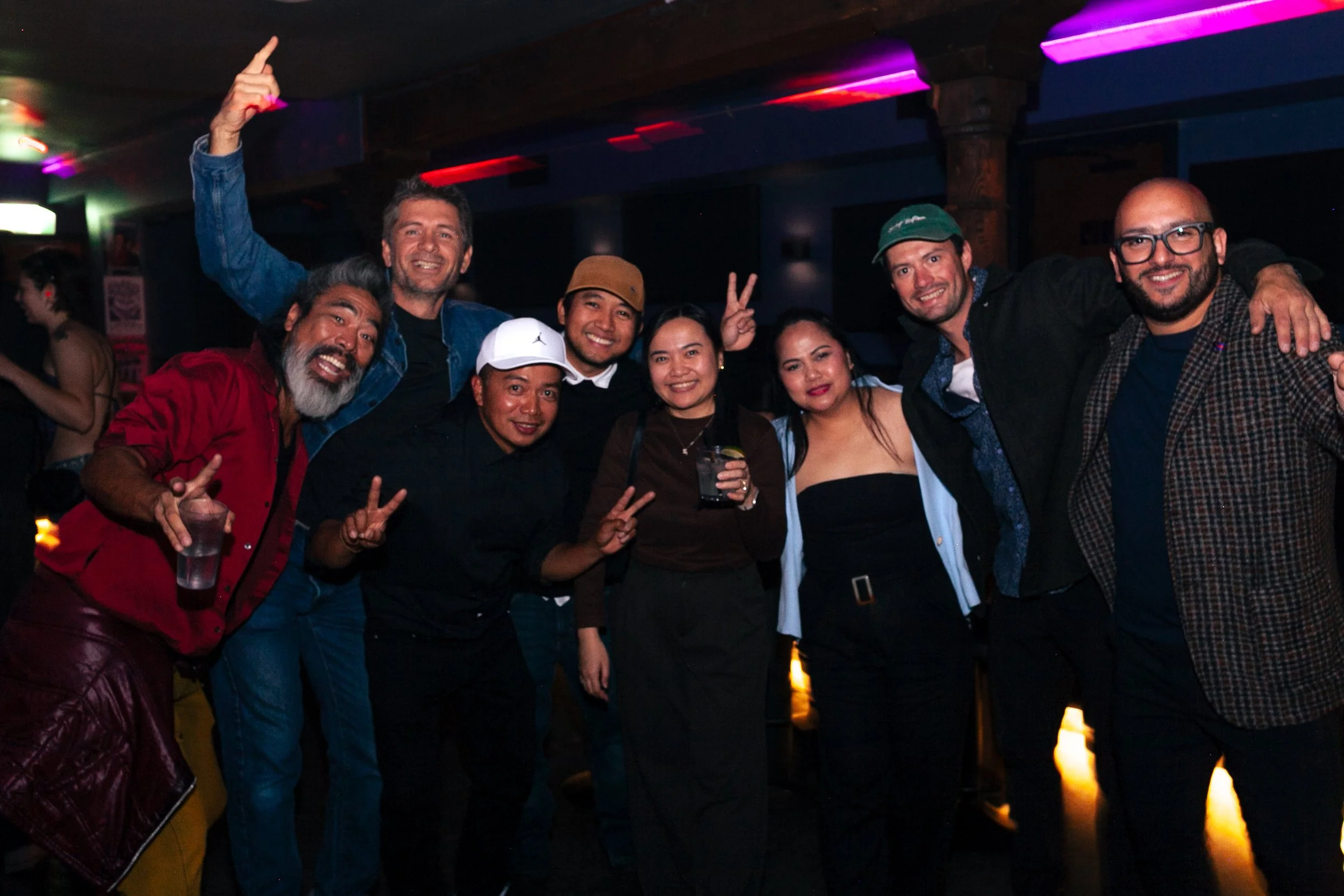 Group of friends at a nightclub smiling and posing for a photo, some making peace signs, with colorful LED lights overhead.