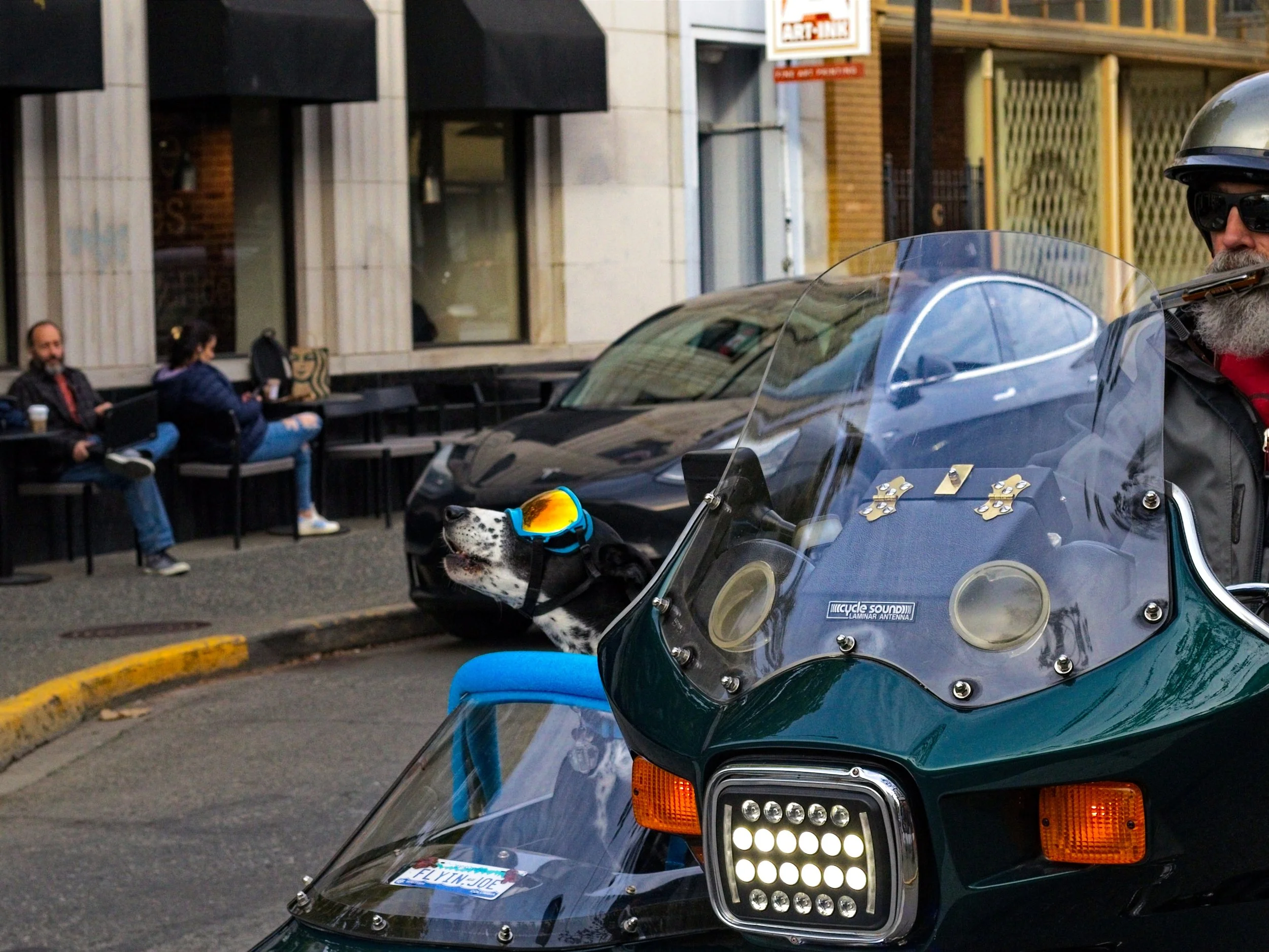 A police officer on a black motorcycle with a clear windshield, wearing a helmet and sunglasses, sitting on the street. In the background, there are two people sitting at a cafe, one with a laptop. The street scene includes buildings with windows and