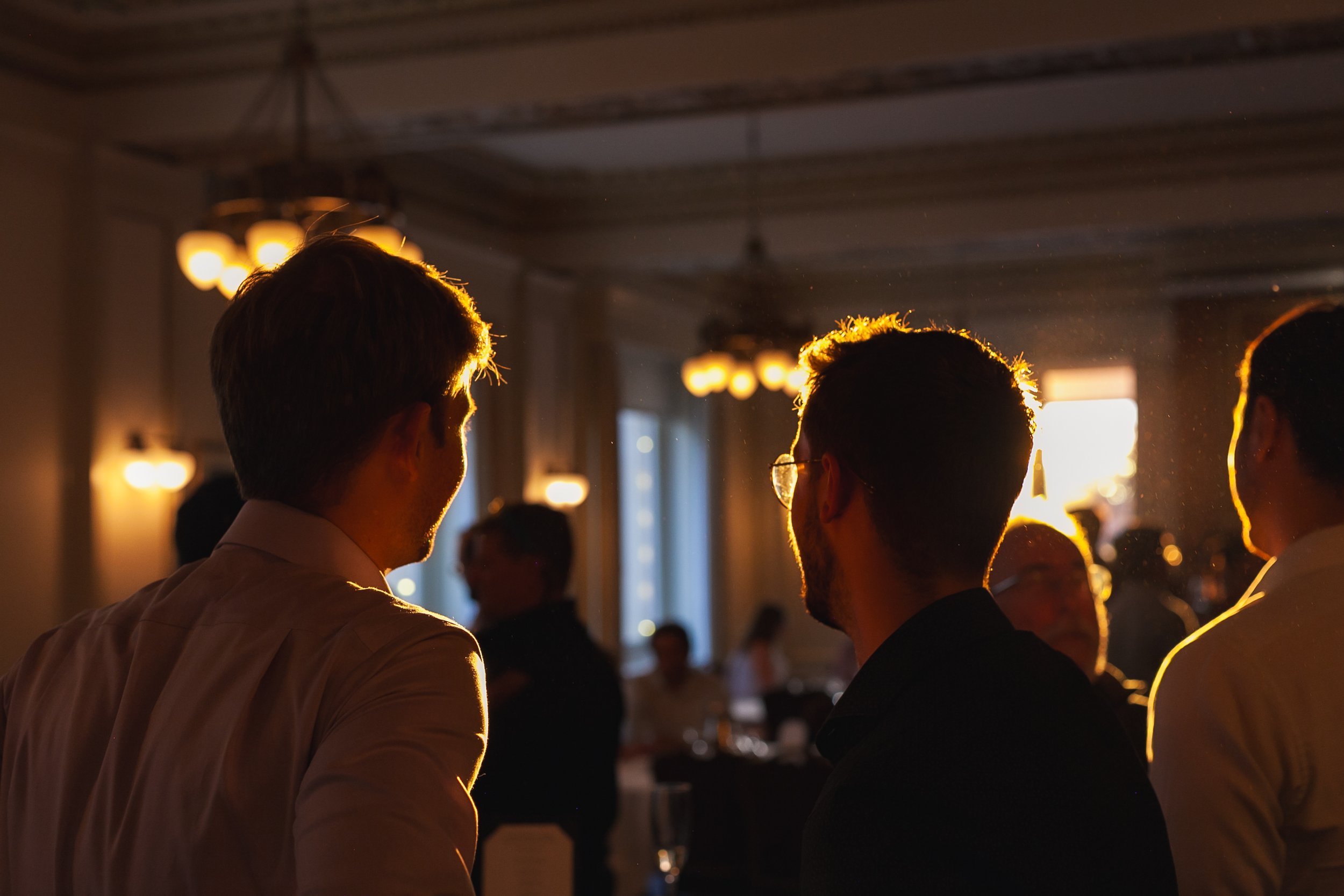 People gathered at an indoor event, talking, with warm lighting and a window in the back.