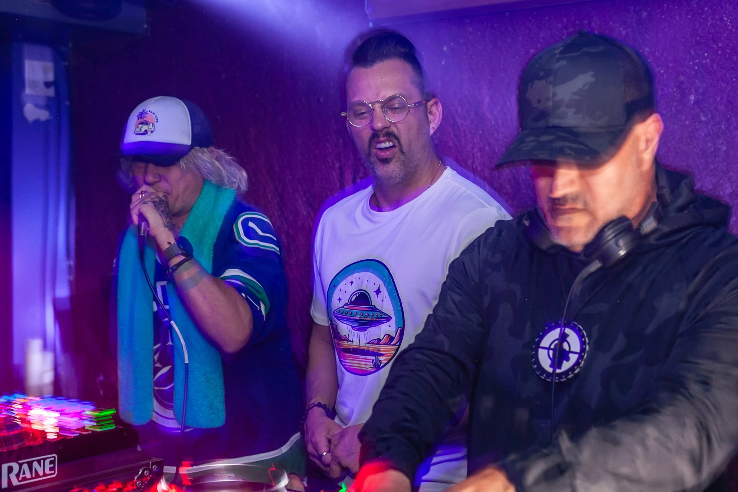Three men standing in a room with purple walls, with one man in the middle wearing glasses and a white t-shirt, and the other two men on either side wearing hats and headphones, possibly DJ equipment visible in the foreground.