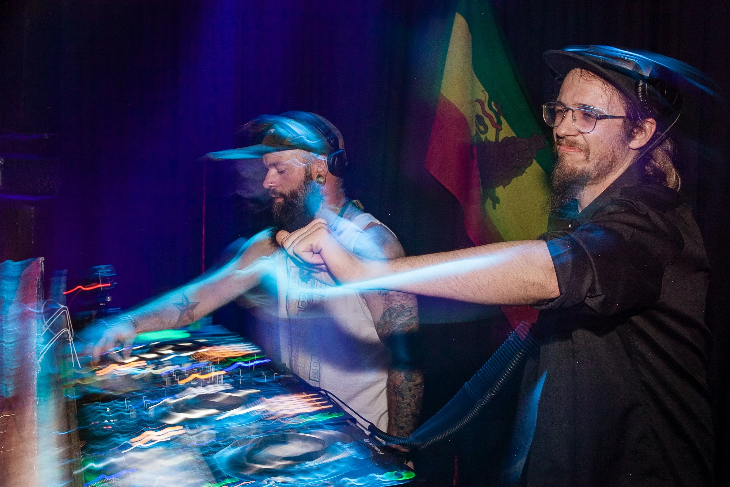 Two men with beards and glasses, wearing helmets, DJing at a nightclub with a Paraguayan flag in the background and colorful light effects.