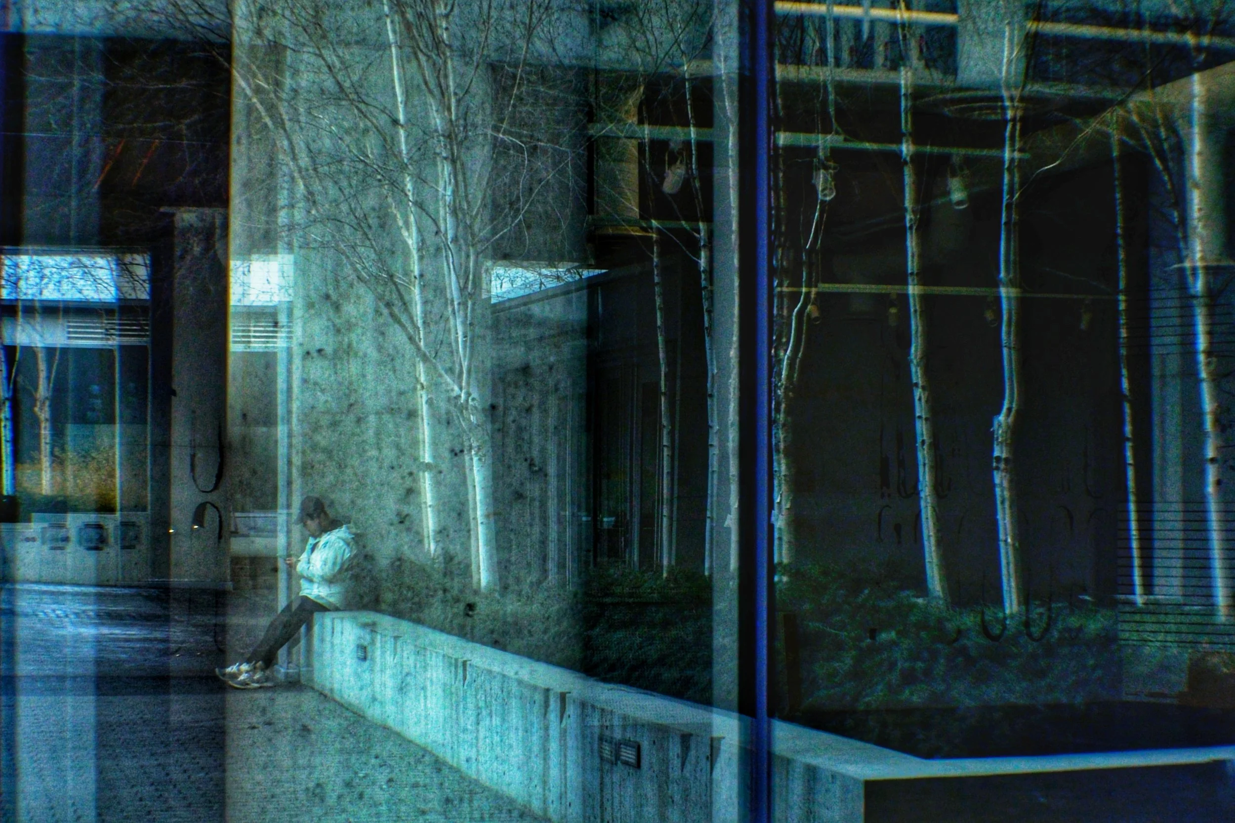 A woman in a white jacket is sitting on a bench in an outdoor area, looking at her phone. There are bare trees and a modern building with glass windows in the background, with reflections visible on the glass.