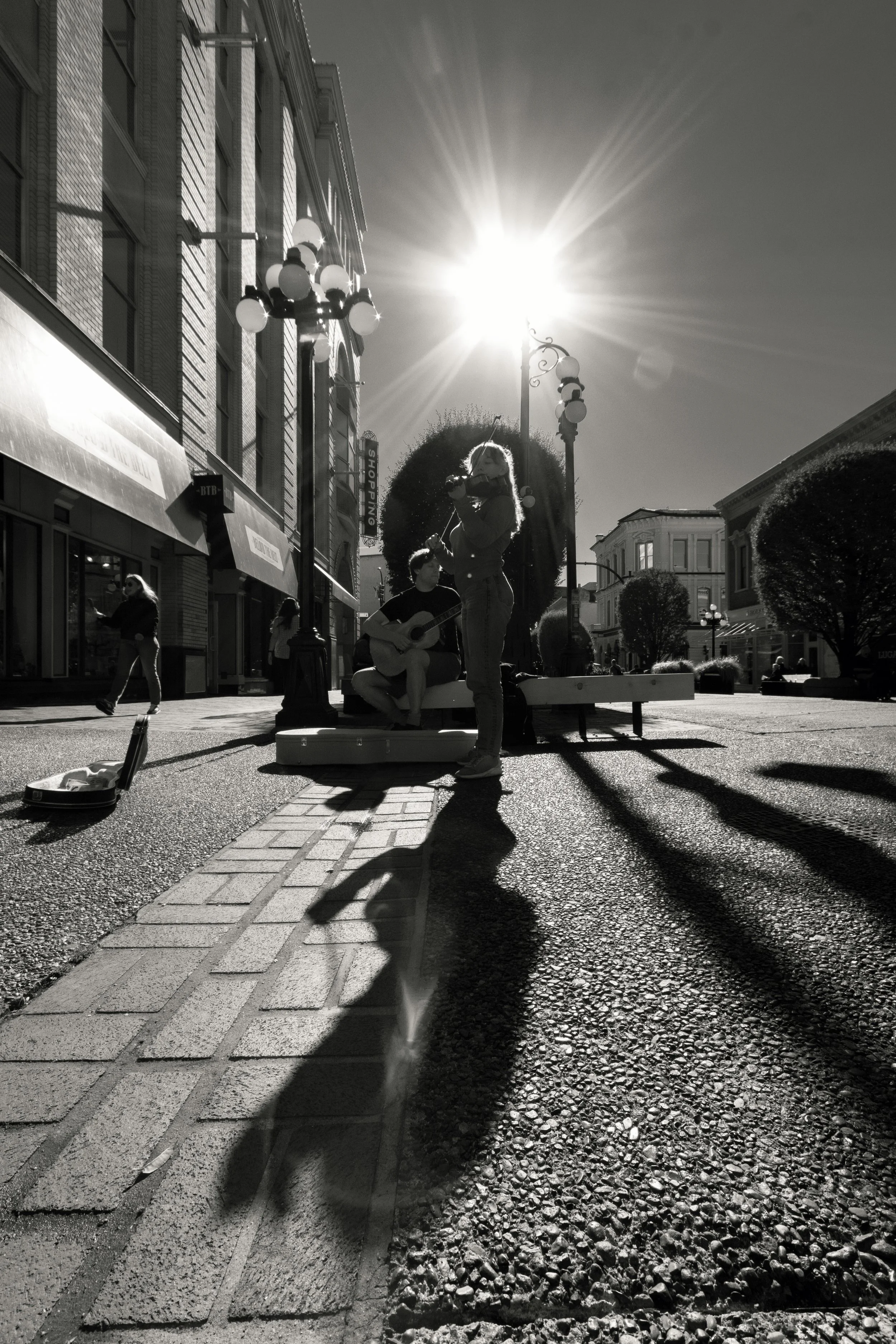 Street scene in black and white with two musicians performing on a bench, a person walking, and a woman standing nearby. Bright sunlight creates shadows and lens flare.