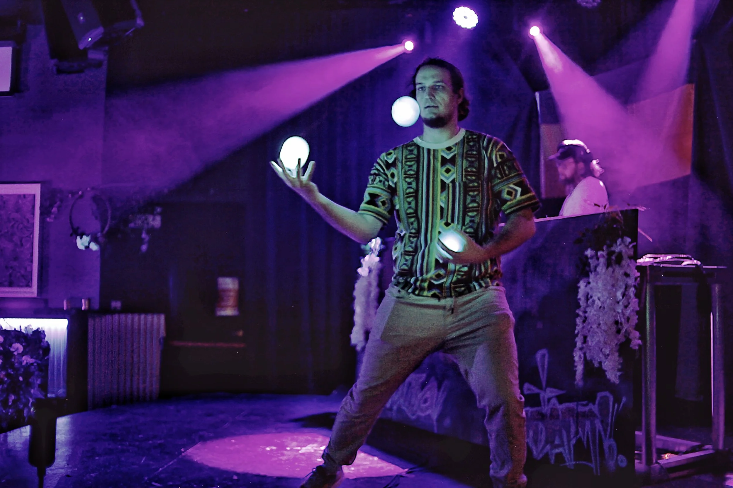 A man with long hair and a beard performing a dance or theatrical act on stage. He is holding a glowing object in his hand, surrounded by purple stage lighting. He is wearing a patterned shirt and light-colored pants.