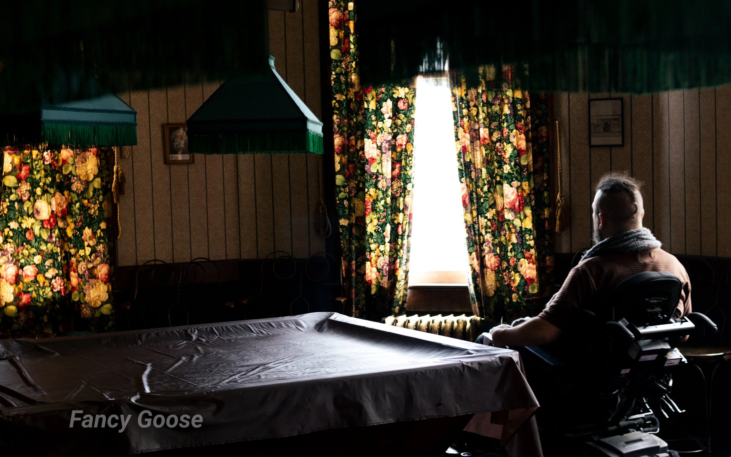A person with a beard in a brown jacket and scarf sitting in a wheelchair in a dimly lit room with floral curtains and green lampshades, with sunlight streaming through a window, and a black tarp covering a surface in the foreground.