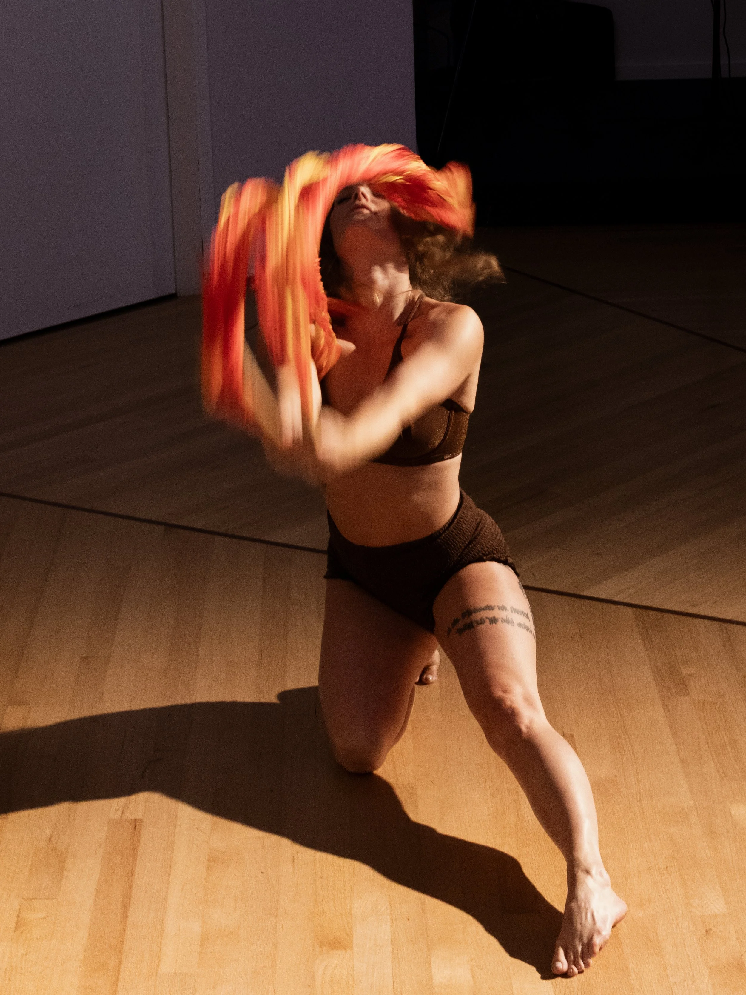 A person performing a dance move on a wooden floor, holding a colorful piece of fabric and balancing on one knee with the other leg extended.