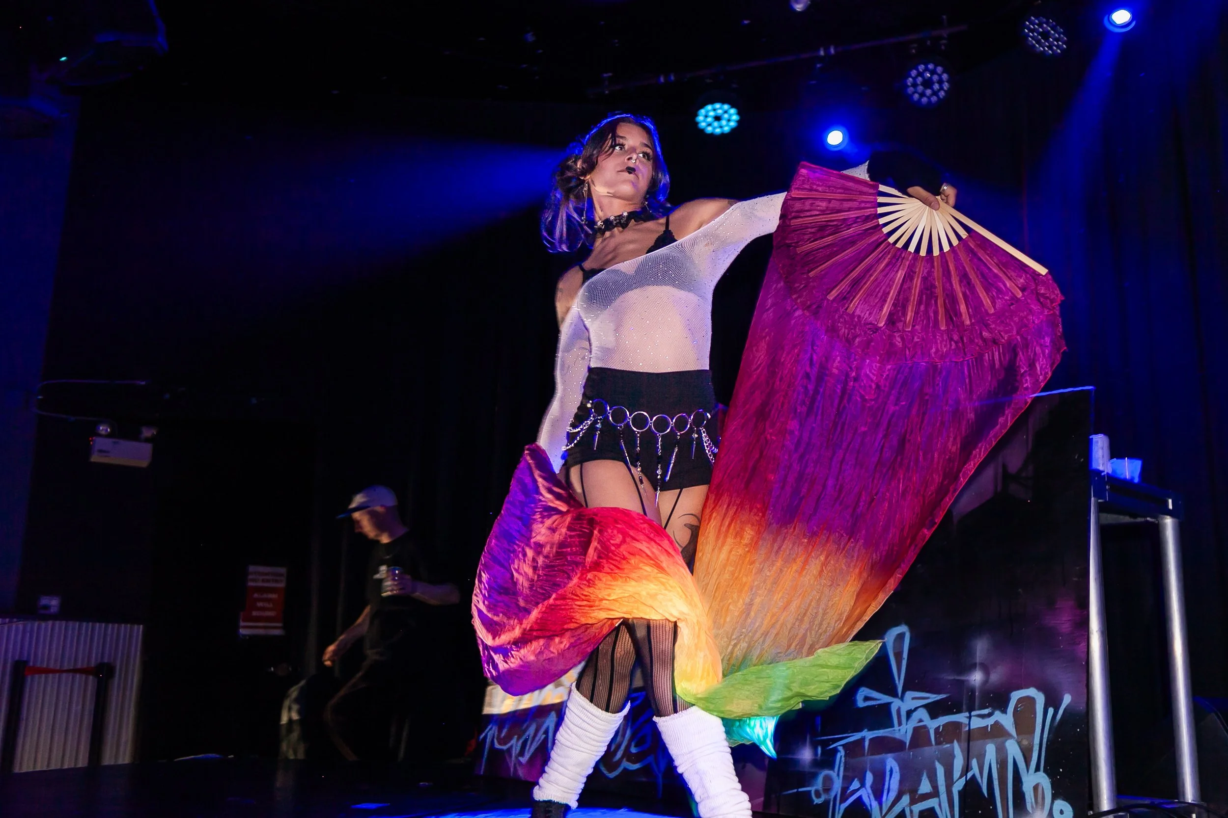 Performer on stage holding a large, colorful paper umbrella with purple, red, orange, yellow, and green sections, dressed in a black and white outfit with fishnet stockings and accessories, under blue stage lighting.