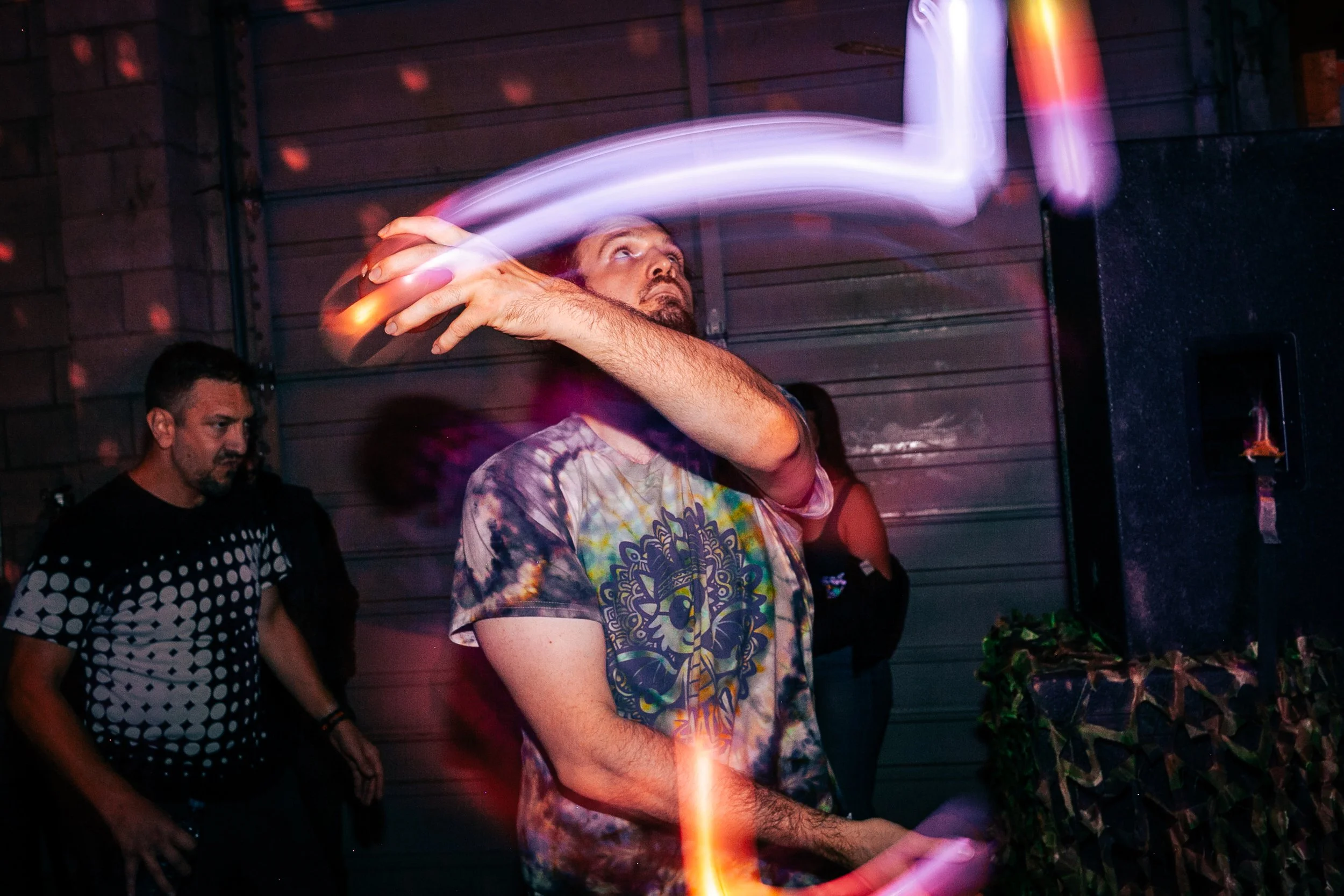 A man with a tattoo sleeve is spinning glow sticks or poi, creating streaks of colorful light in a dark indoor setting. Another man with a beard, wearing a black and white patterned shirt, stands nearby watching. The background includes a wooden wall