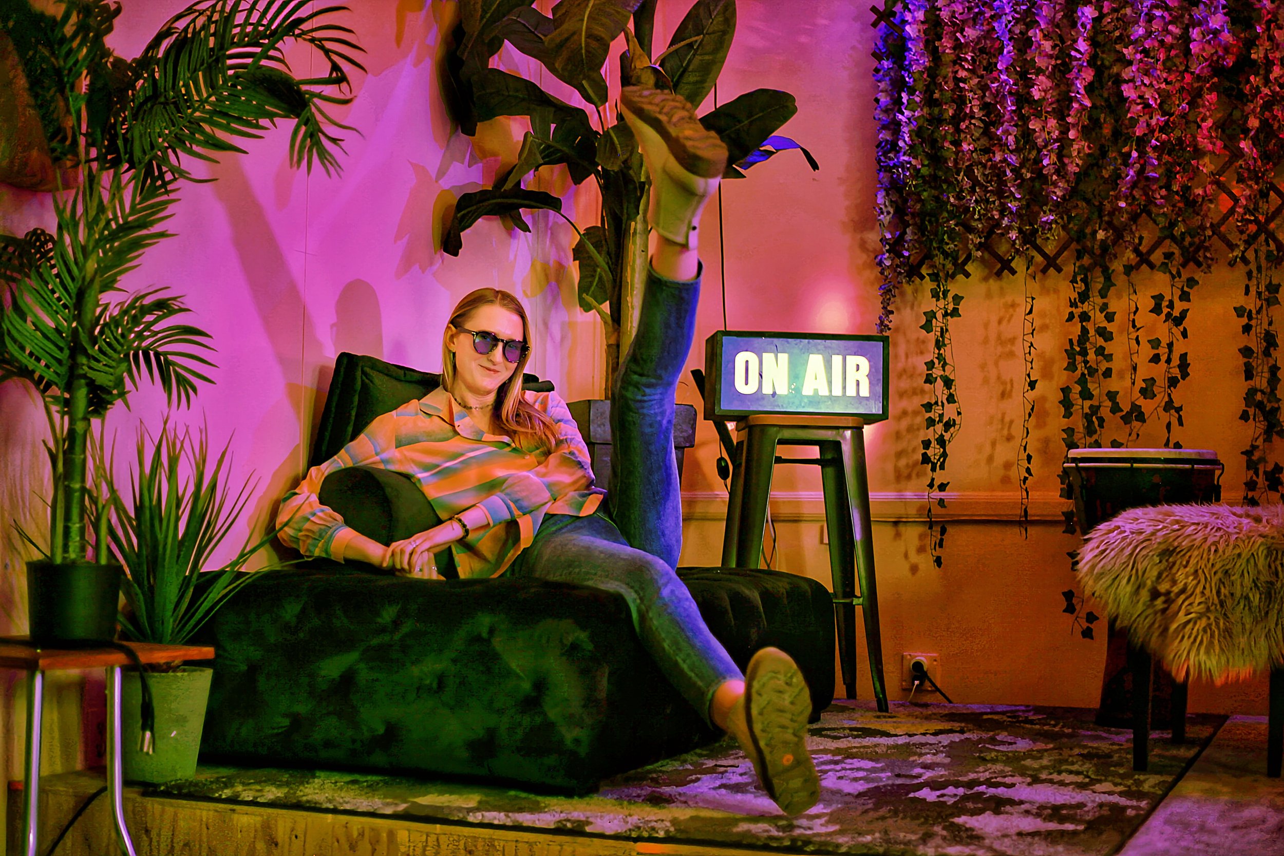 A woman with long blonde hair, wearing glasses, a plaid shirt, and jeans, is lounging on a black sofa in a room with vibrant pink and purple lighting. She seems relaxed, with one leg crossed over the other and her arm resting on her lap. The background features a large potted plant, a small TV with an "On Air" sign, and hanging ivy on the wall, creating a cozy, bohemian atmosphere.